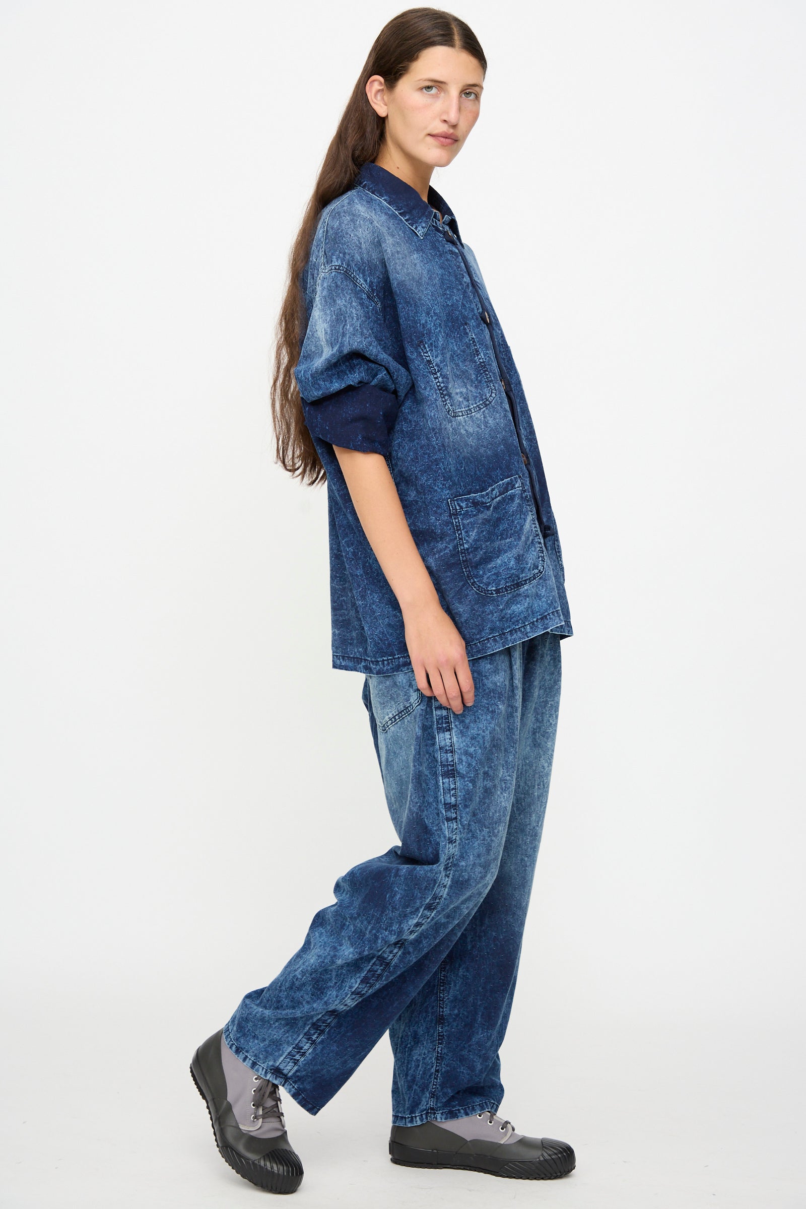 A person with long brown hair wears a loose-fitting, dark blue denim jacket and Dr. Collectors 6 oz. Cotton P43 Kyoto Pant in Sunfaded Indigo with gray sneakers, standing against a plain white background.