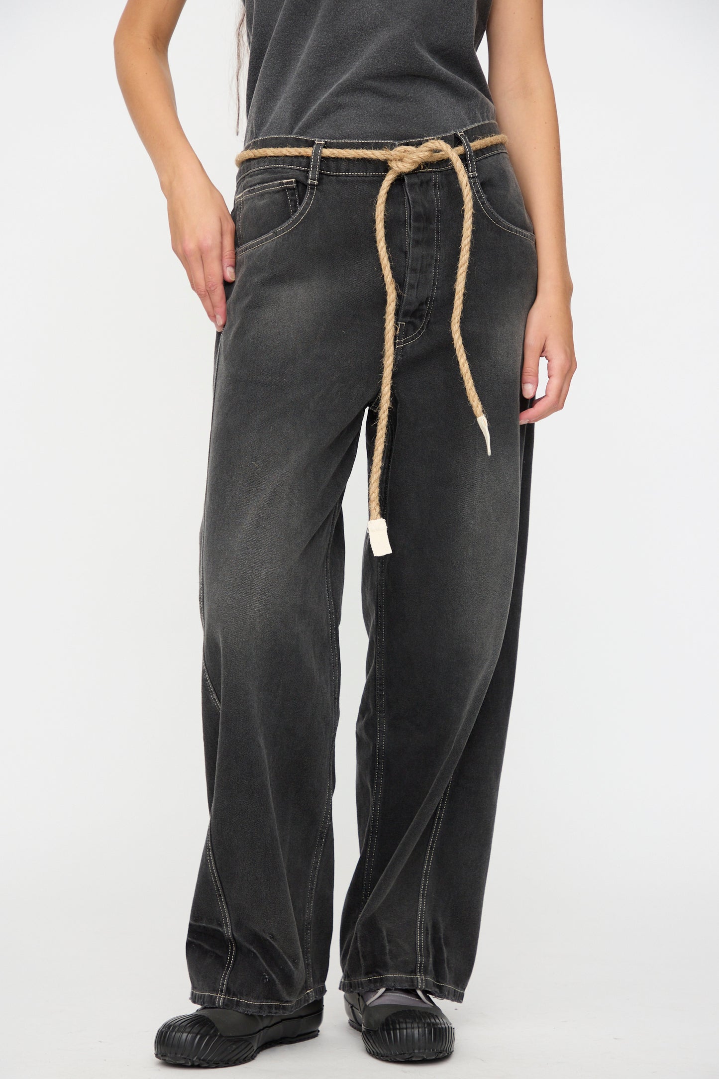 A person wearing the Dr. Collectors 9 oz. Denim P84 Dungaree Pant in Sunfaded Black, styled with a braided hemp rope belt, black shoes, and a dark top, stands against a plain white background.