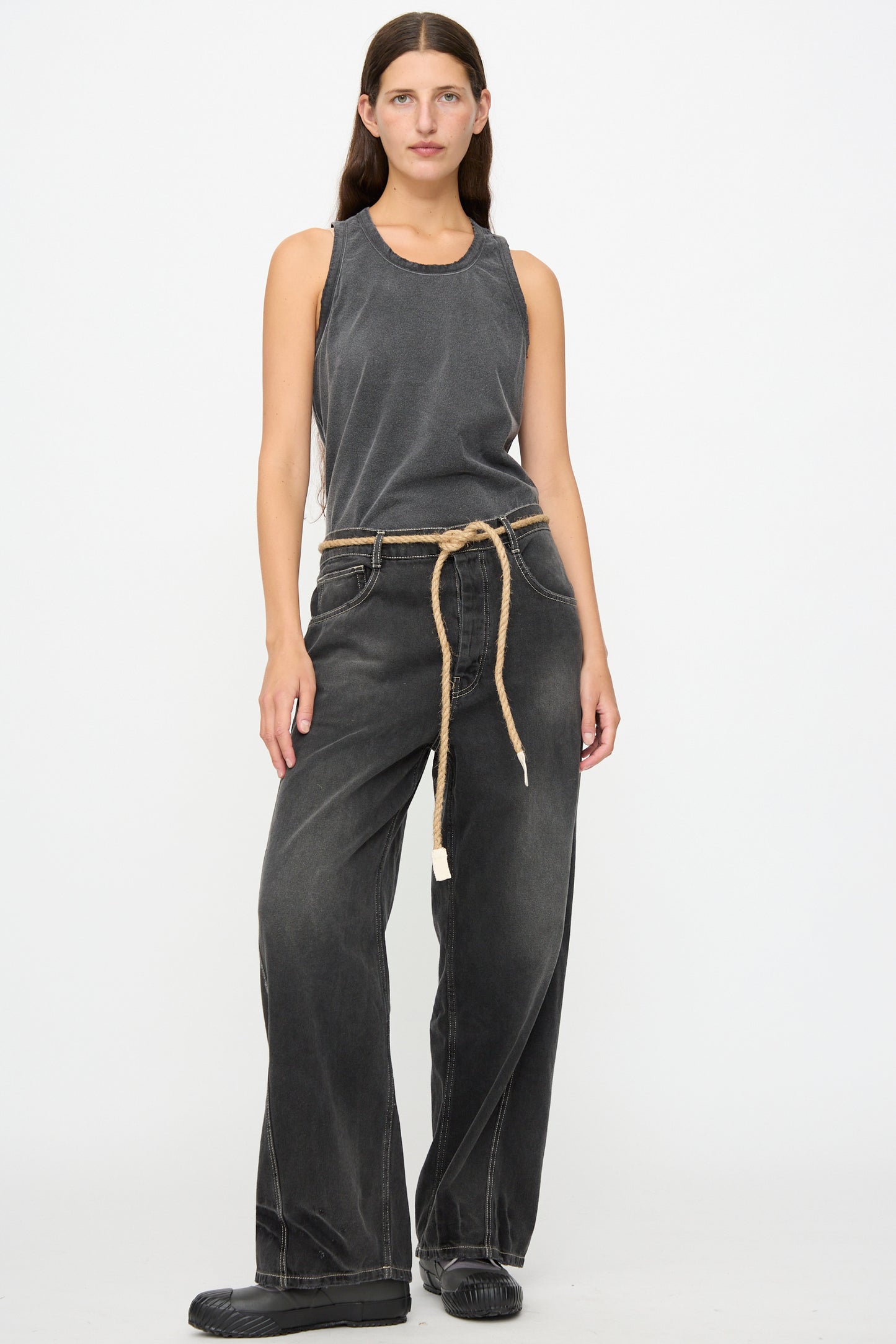 A person wearing a gray tank top, Dr. Collectors 9 oz. Denim P84 Dungaree Pant in Sunfaded Black with a braided hemp rope belt, and black shoes stands against a plain white background.