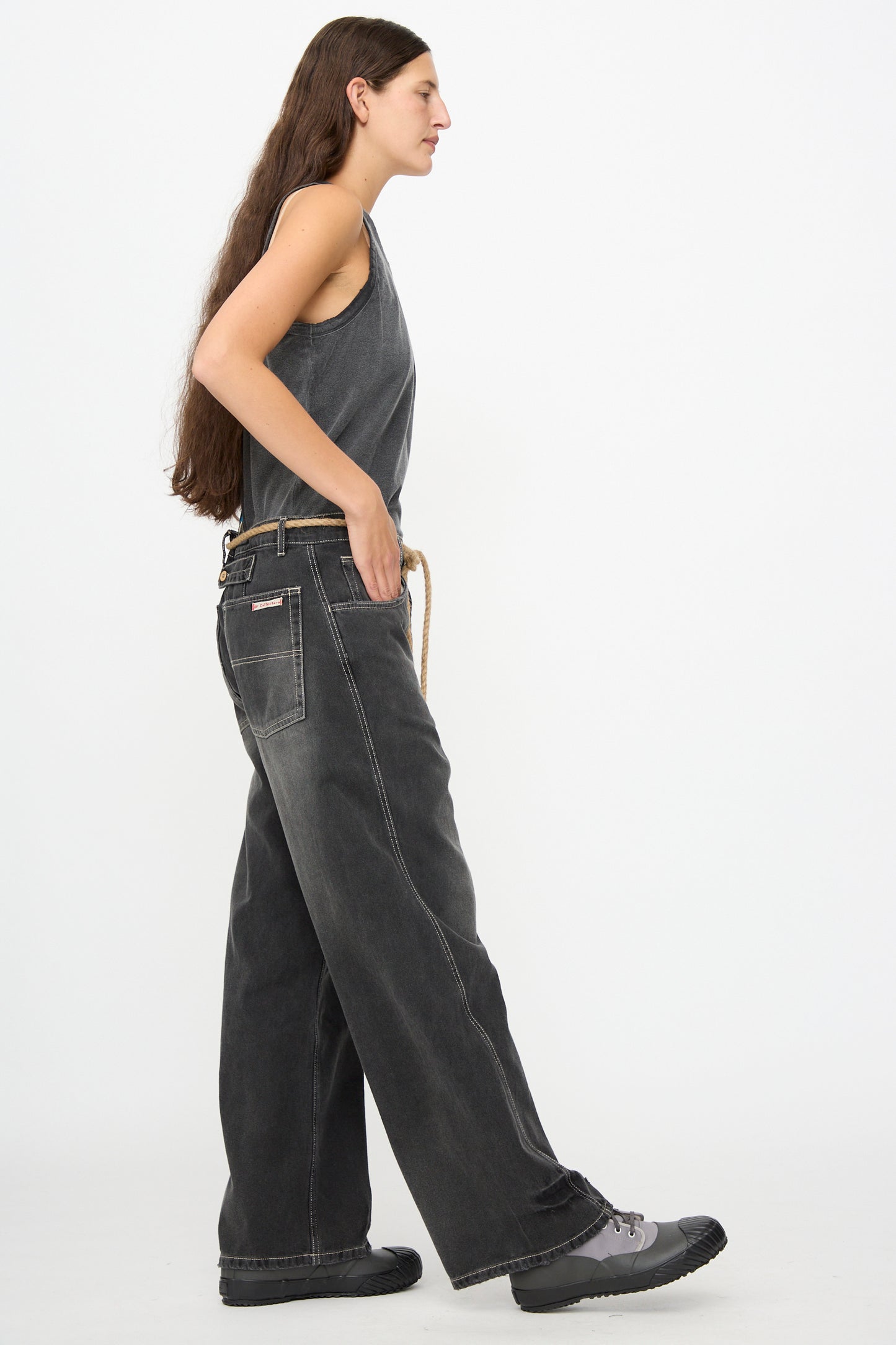 A person with long brown hair wearing a sleeveless gray top, Dr. Collectors 9 oz. Denim P84 Dungaree Pant in Sunfaded Black featuring wide legs and visible stitching, and black shoes stands in profile against a plain white background.