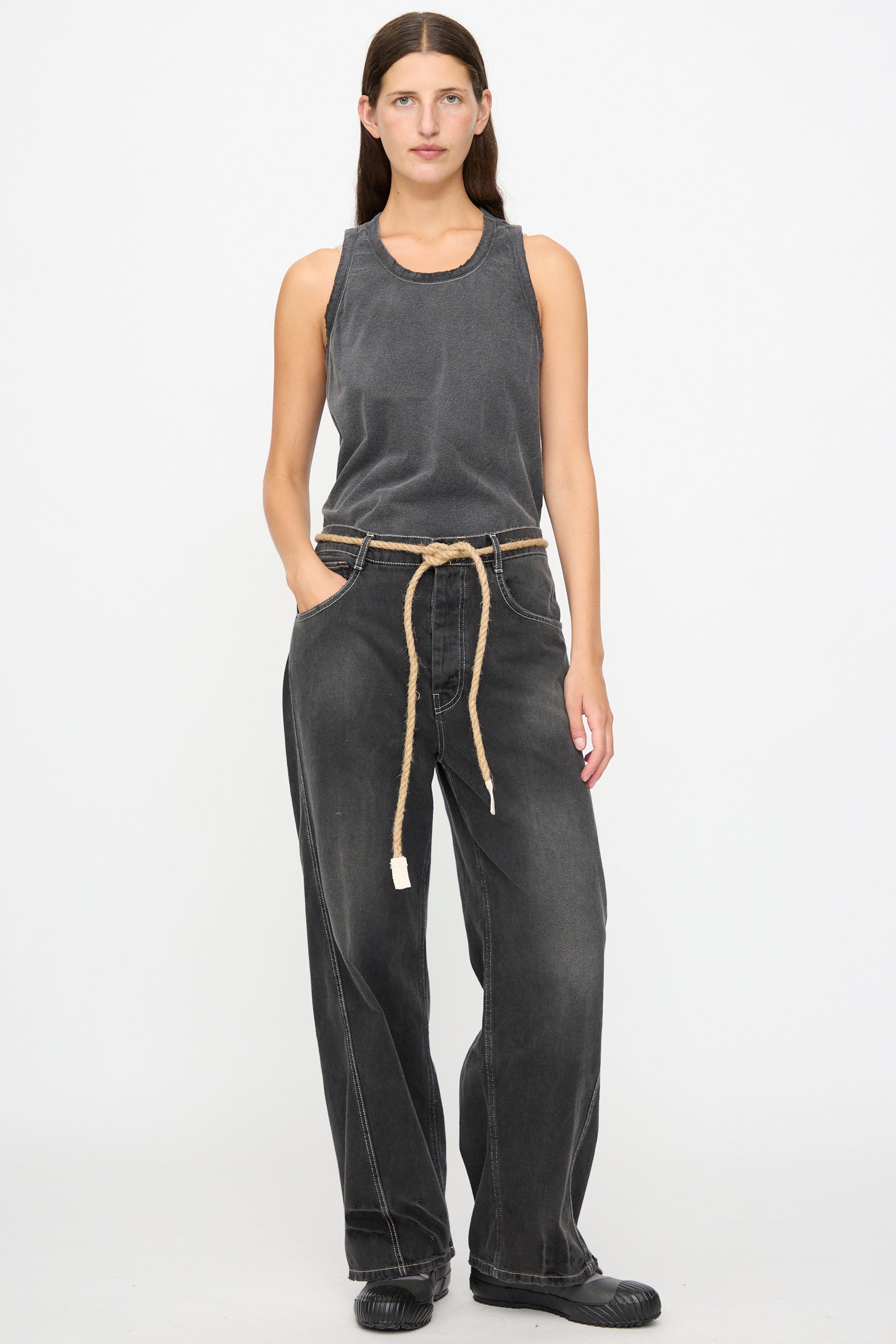 A person with long brown hair wears a faded black sleeveless top, Dr. Collectors 9 oz. Denim P84 Dungaree Pant in Sunfaded Black styled with a braided hemp rope belt, and black shoes, standing against a plain white background.