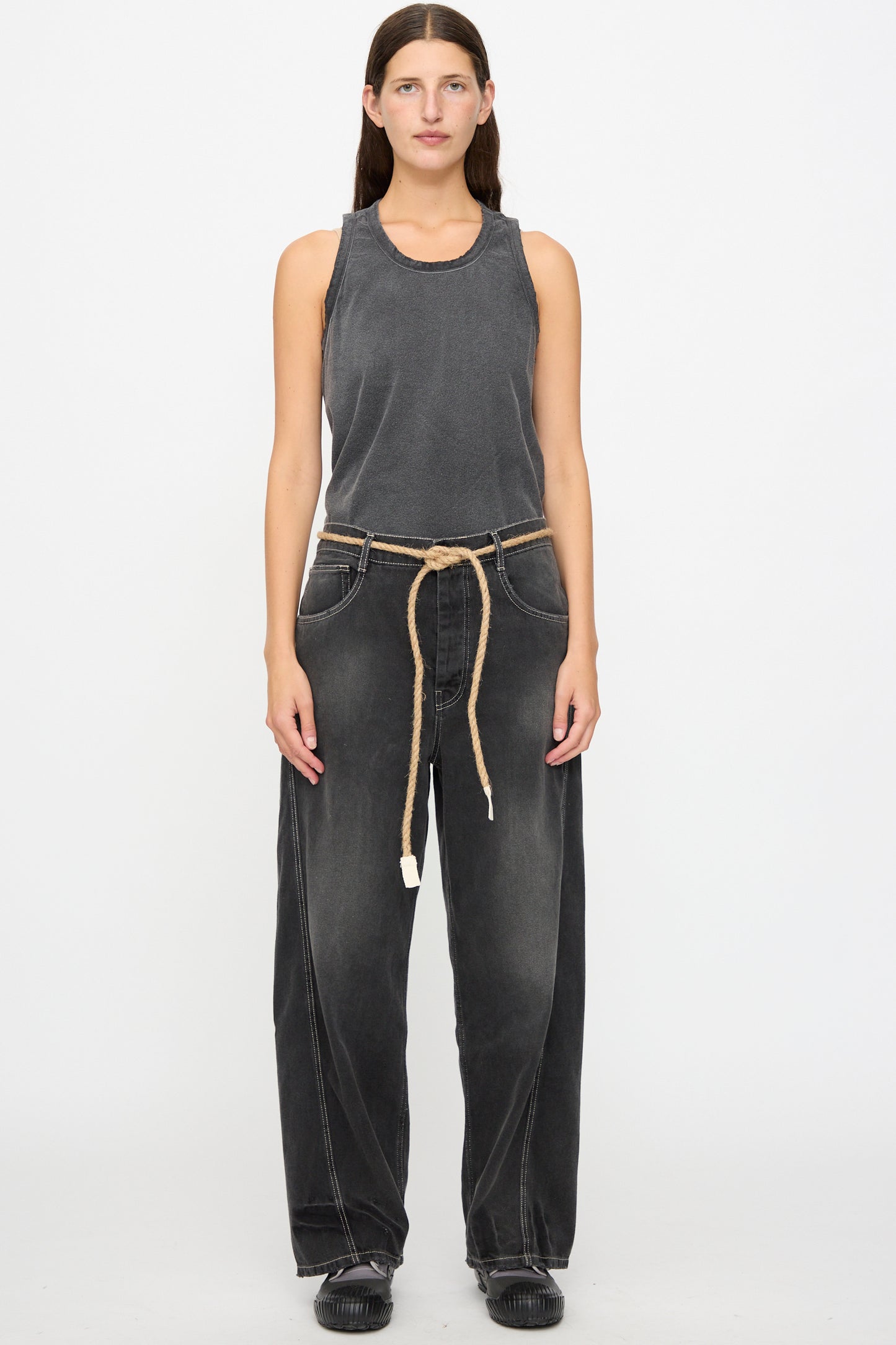 A person stands facing forward against a plain white background, wearing a sleeveless gray top, the Dr. Collectors 9 oz. Denim P84 Dungaree Pant in Sunfaded Black with a braided hemp rope belt, and black shoes.