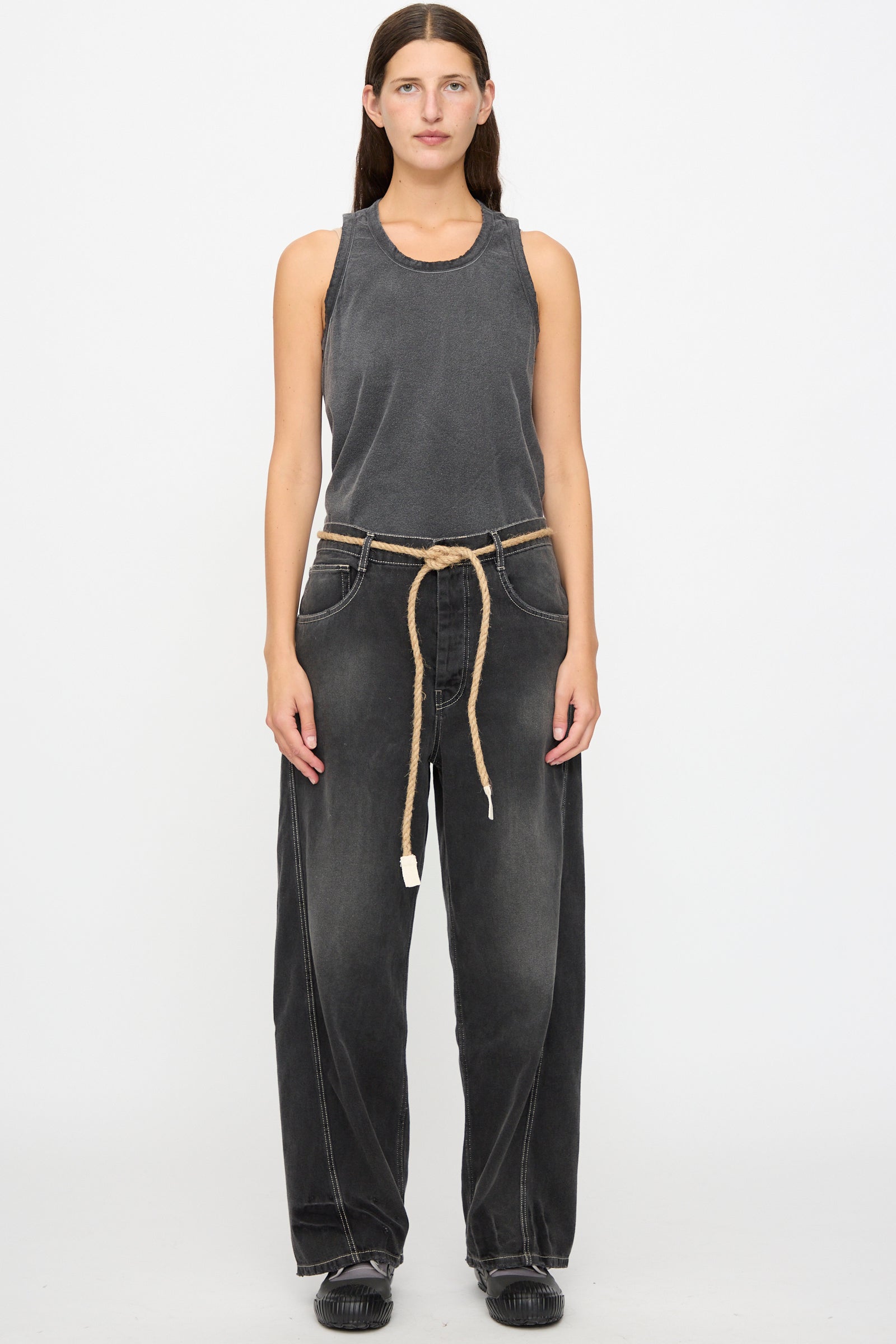 A person stands facing forward against a plain white background, wearing a sleeveless gray top, the Dr. Collectors 9 oz. Denim P84 Dungaree Pant in Sunfaded Black with a braided hemp rope belt, and black shoes.