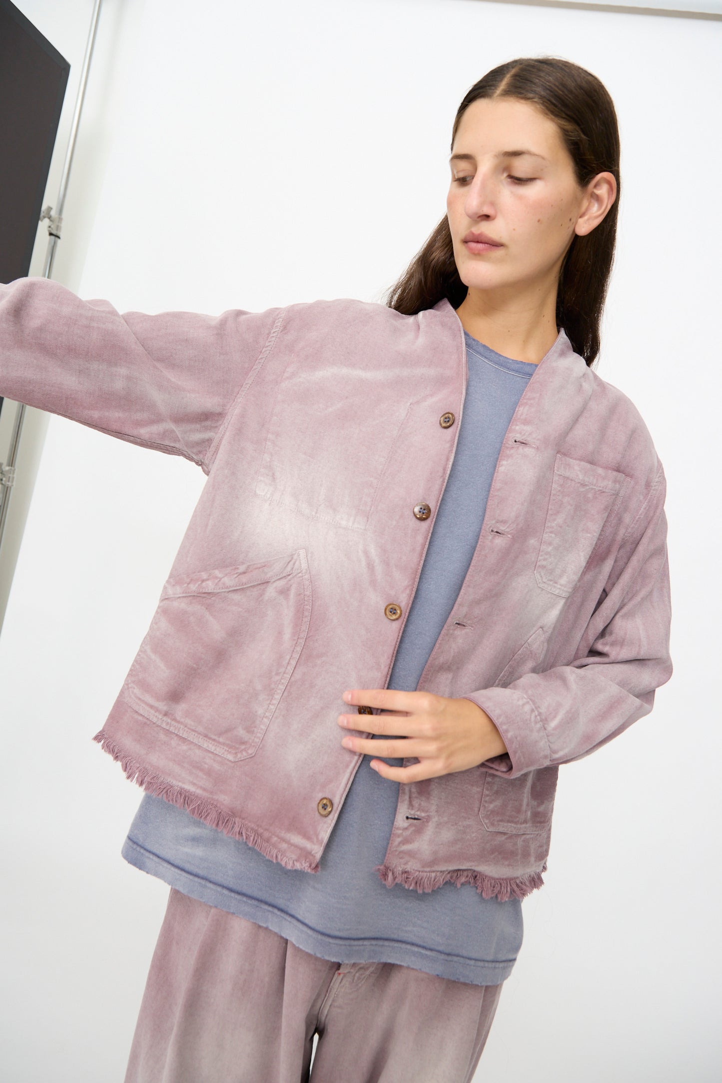 A person wearing the Dr. Collectors 9 oz. Okinawa Jacket in Sunfaded Dusty Orchid—a mauve vintage-inspired piece with frayed edges, crafted from an organic cotton blend—layered over a blue shirt and matching mauve pants, stands indoors with one arm extended to the side.