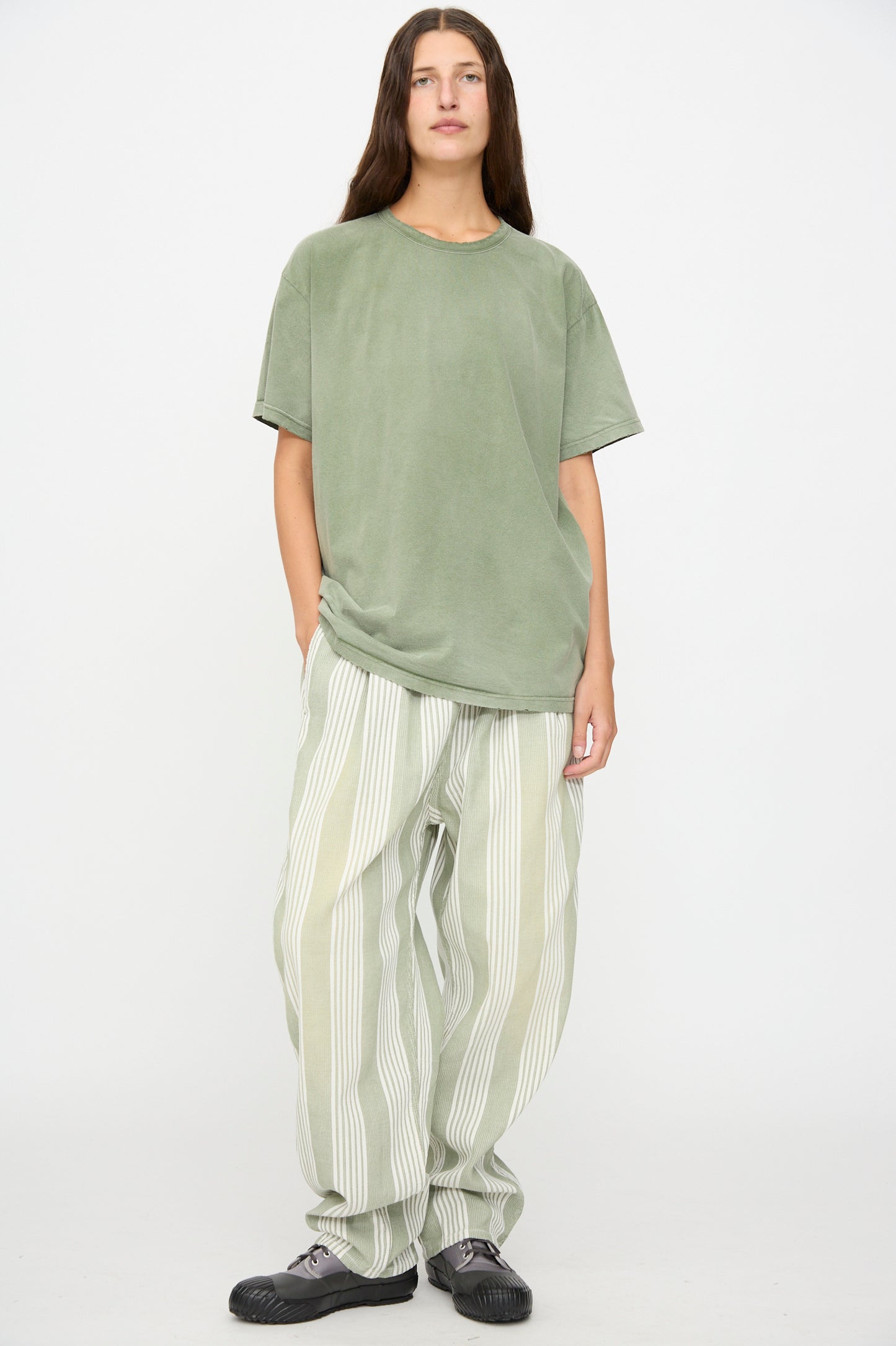 A person stands against a plain background wearing a loose green T-shirt and Dr. Collectors 9 oz. P43 Kyoto Multi Stripe Pant in Sunfaded Light Green—wide, relaxed cotton pants with light green and white stripes, featuring an elastic waistband and made in California. Black shoes complete the look, with hands in pockets.