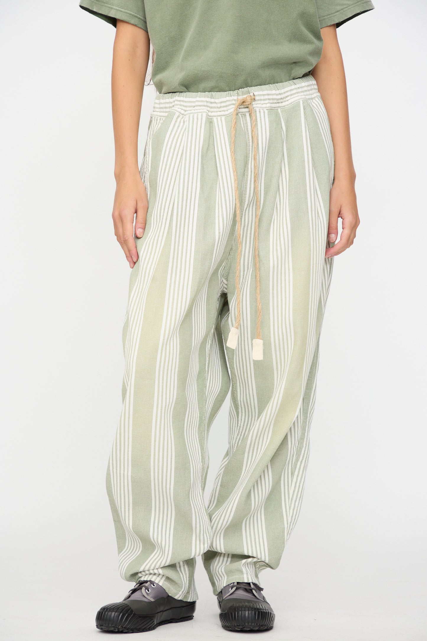 A person is wearing Dr. Collectors’ 9 oz. P43 Kyoto Multi Stripe Pant in Sunfaded Light Green—relaxed cotton pants featuring an elastic waistband and drawstring—paired with a green short-sleeve shirt and dark green shoes, standing against a plain white background.