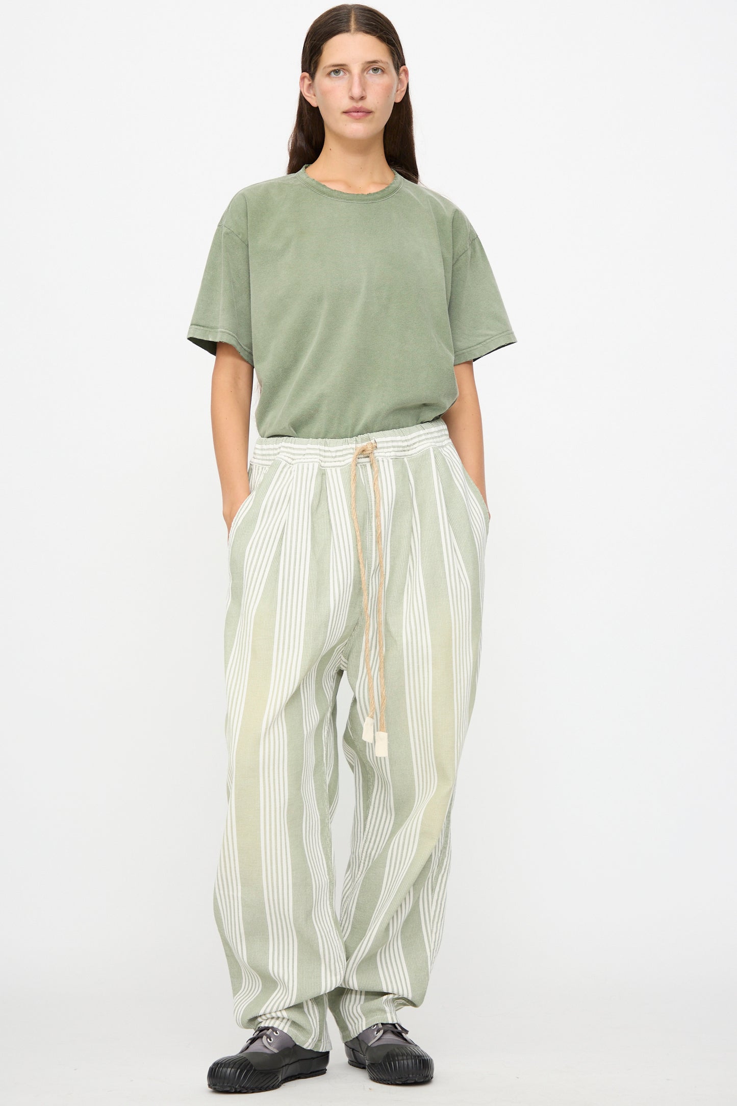 A person stands against a white background, wearing a sage green t-shirt and the Dr. Collectors 9 oz. P43 Kyoto Multi Stripe Pant in Sunfaded Light Green—relaxed cotton pants featuring light green and white stripes, an elastic waistband with drawstring, and made in California. Their hands are in the pockets, paired with black shoes.