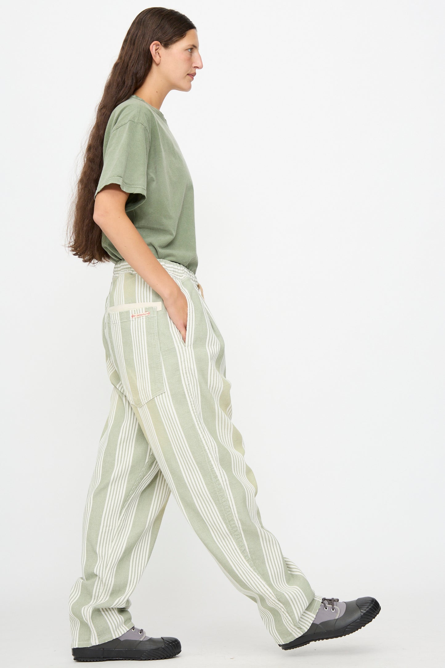 A person in profile view wearing a green t-shirt, Dr. Collectors 9 oz. P43 Kyoto Multi Stripe Pant in Sunfaded Light Green with an elastic waistband, and gray shoes stands against a plain white background with hands in pockets.