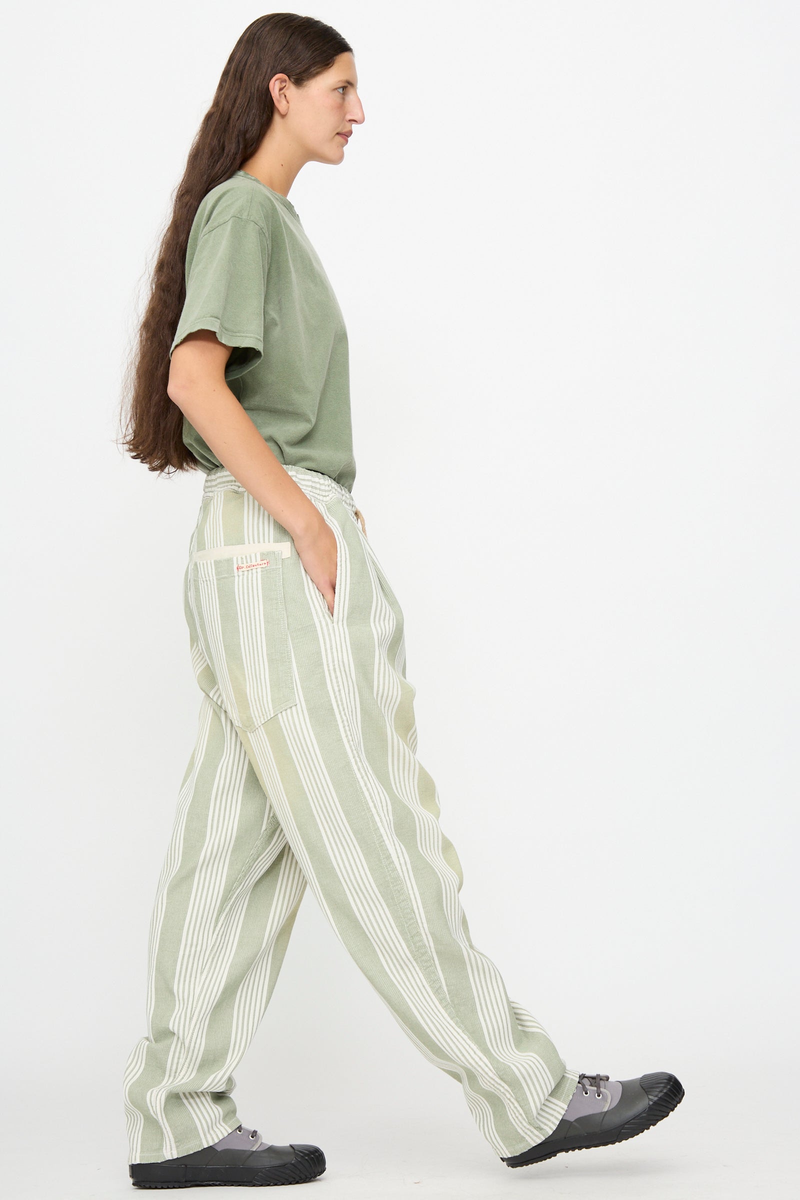 A person in profile view wearing a green t-shirt, Dr. Collectors 9 oz. P43 Kyoto Multi Stripe Pant in Sunfaded Light Green with an elastic waistband, and gray shoes stands against a plain white background with hands in pockets.