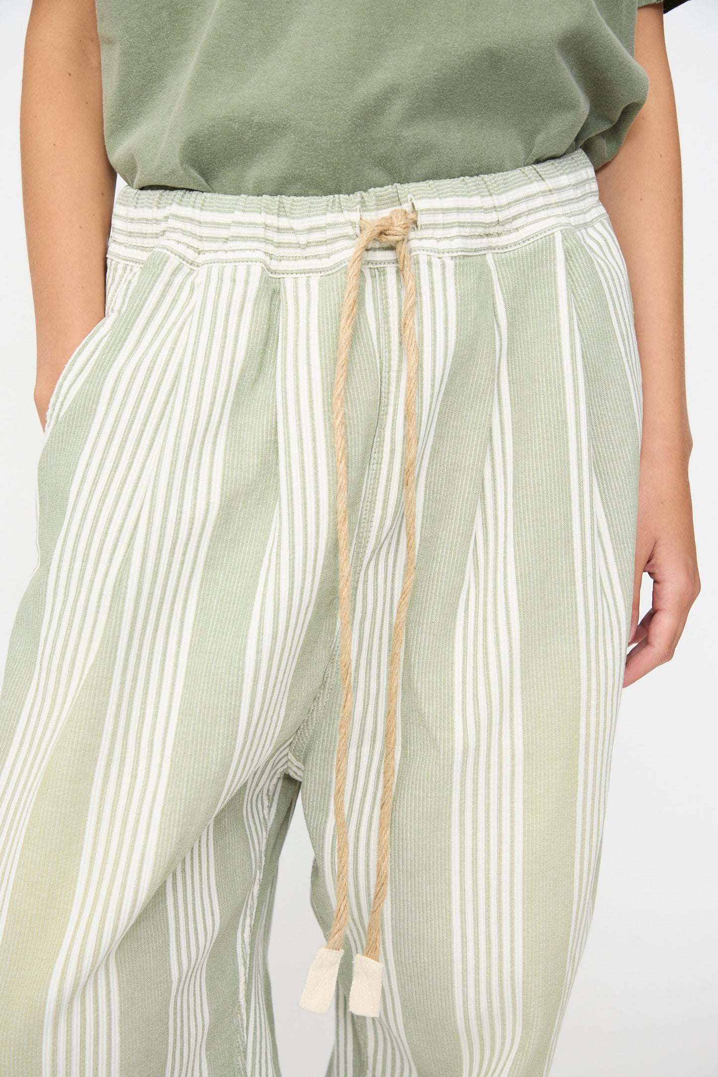 A person wears the Dr. Collectors 9 oz. P43 Kyoto Multi Stripe Pant in Sunfaded Light Green—a relaxed, cotton pant featuring green and white vertical stripes, a beige drawstring, and a comfortable elastic waistband—styled with a green shirt and one hand in their pocket. Thoughtfully made in California.