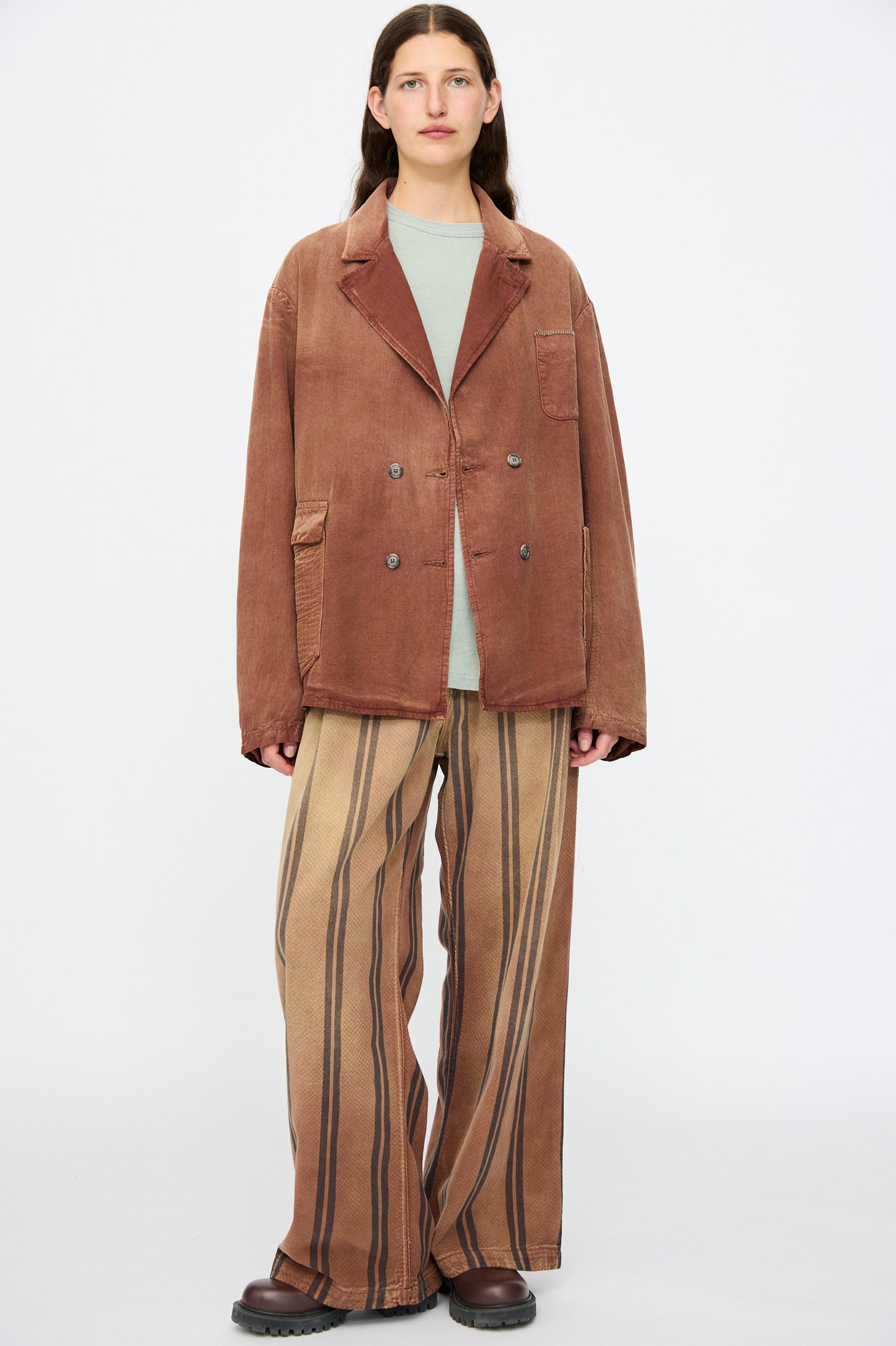 A person stands against a white background wearing the Dr. Collectors Bogart Jacket in Nuts, a loose vintage-style brown jacket, paired with a light green shirt, wide striped brown pants, and dark brown shoes.