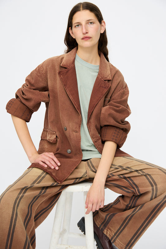 A person sits on a white stool wearing the Dr. Collectors Bogart Jacket in Nuts, a brown corduroy blazer with vintage appeal, paired with a sage green t-shirt and wide-leg brown striped pants against a plain light background.