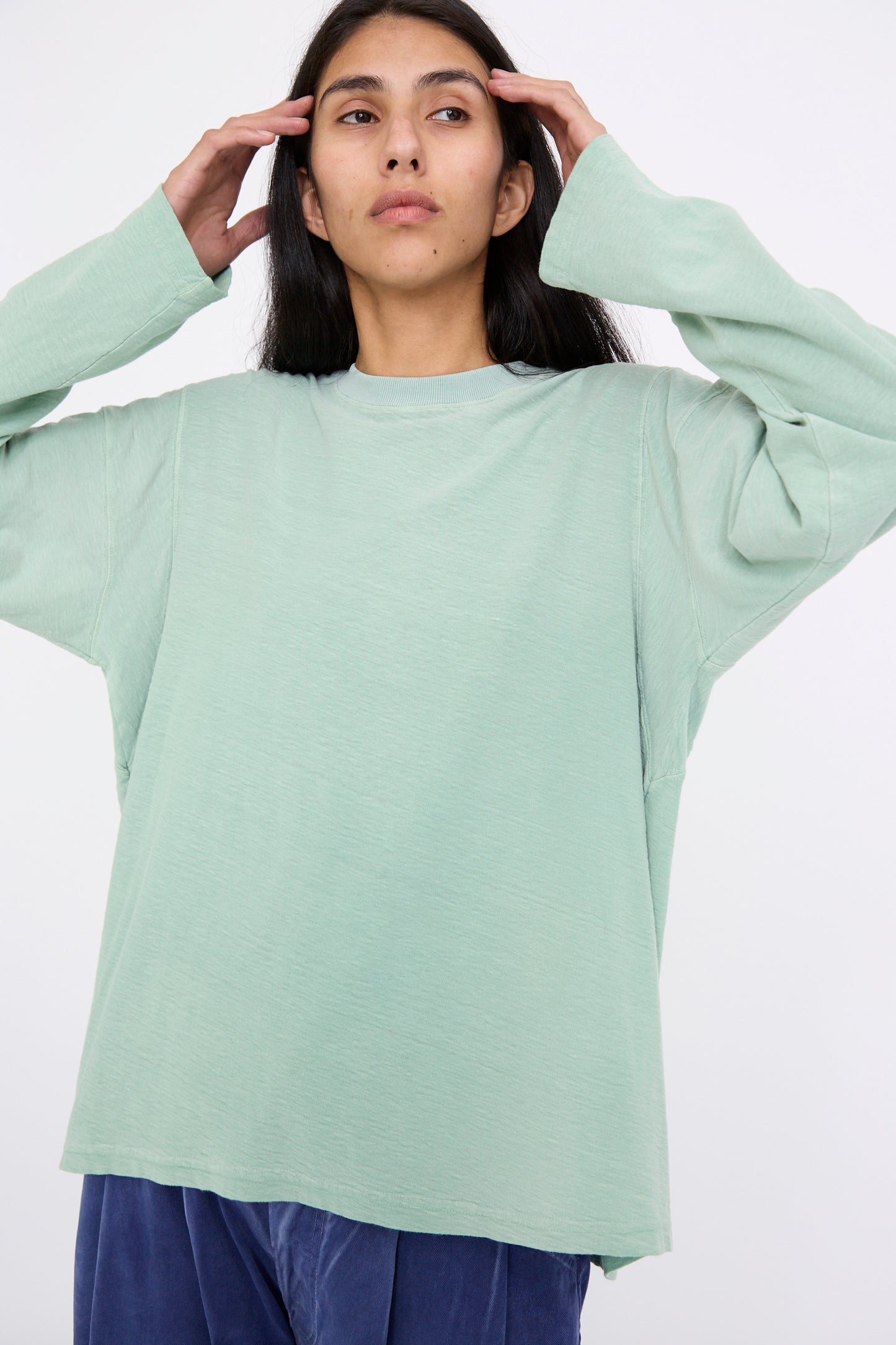 A person wearing the Cotton Derrick Tee in Sunfaded Smoke Green by Dr. Collectors and blue pants, with their hands near their head, looking upwards.