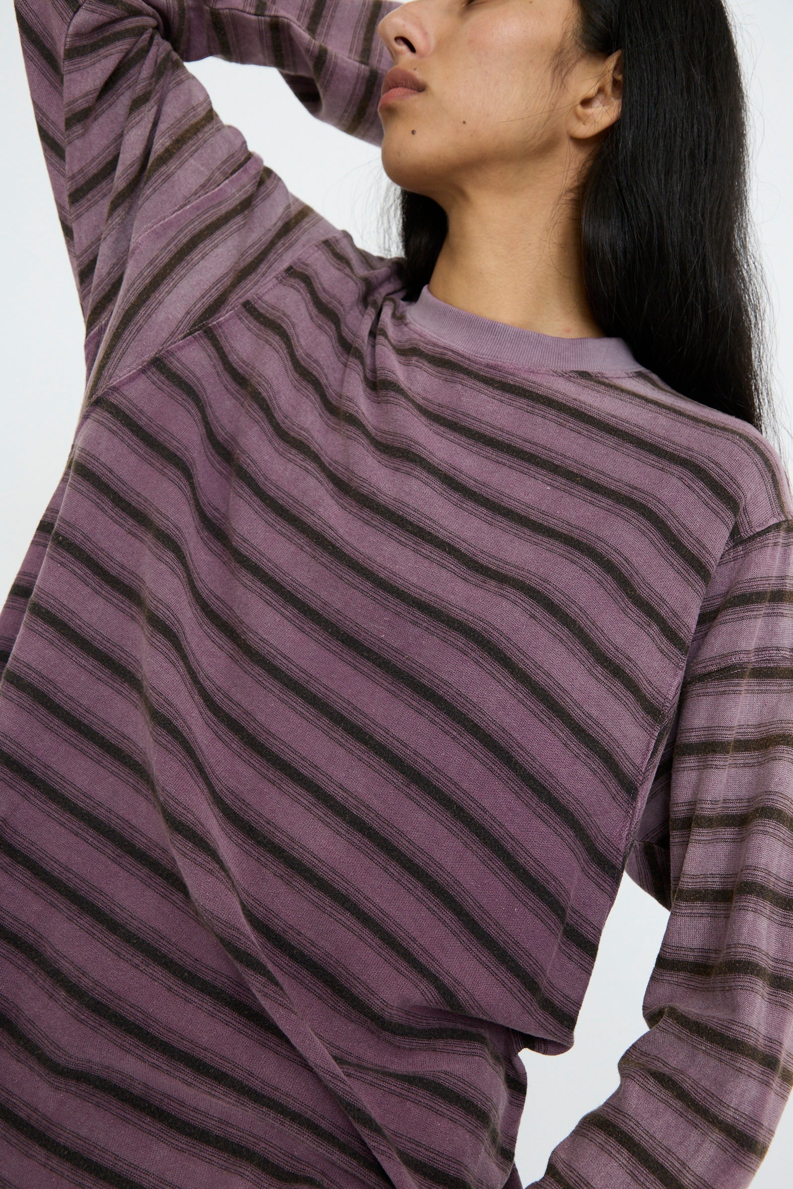 A person wearing the Dr. Collectors Hemp and Cotton Derrick Tee Stripes in Sunfaded Mauve, featuring purple and black stripes and long sleeves, poses with one arm bent and a hand behind their head against a white background. 