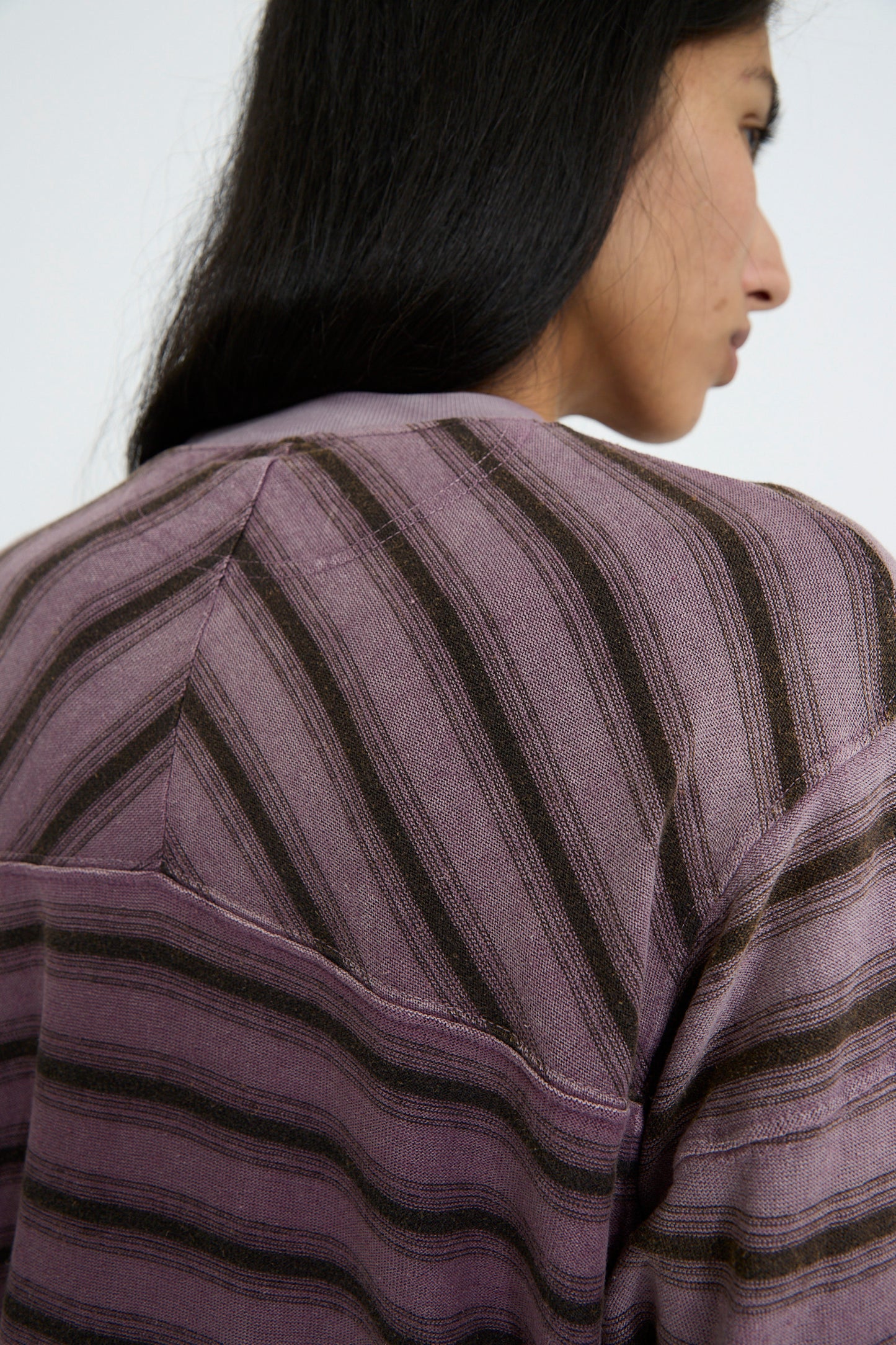 Close-up of a person with long dark hair wearing the Hemp and Cotton Derrick Tee Stripes in Sunfaded Mauve by Dr. Collectors, featuring purple and brown stripes and crafted from recycled cotton, viewed from the back.