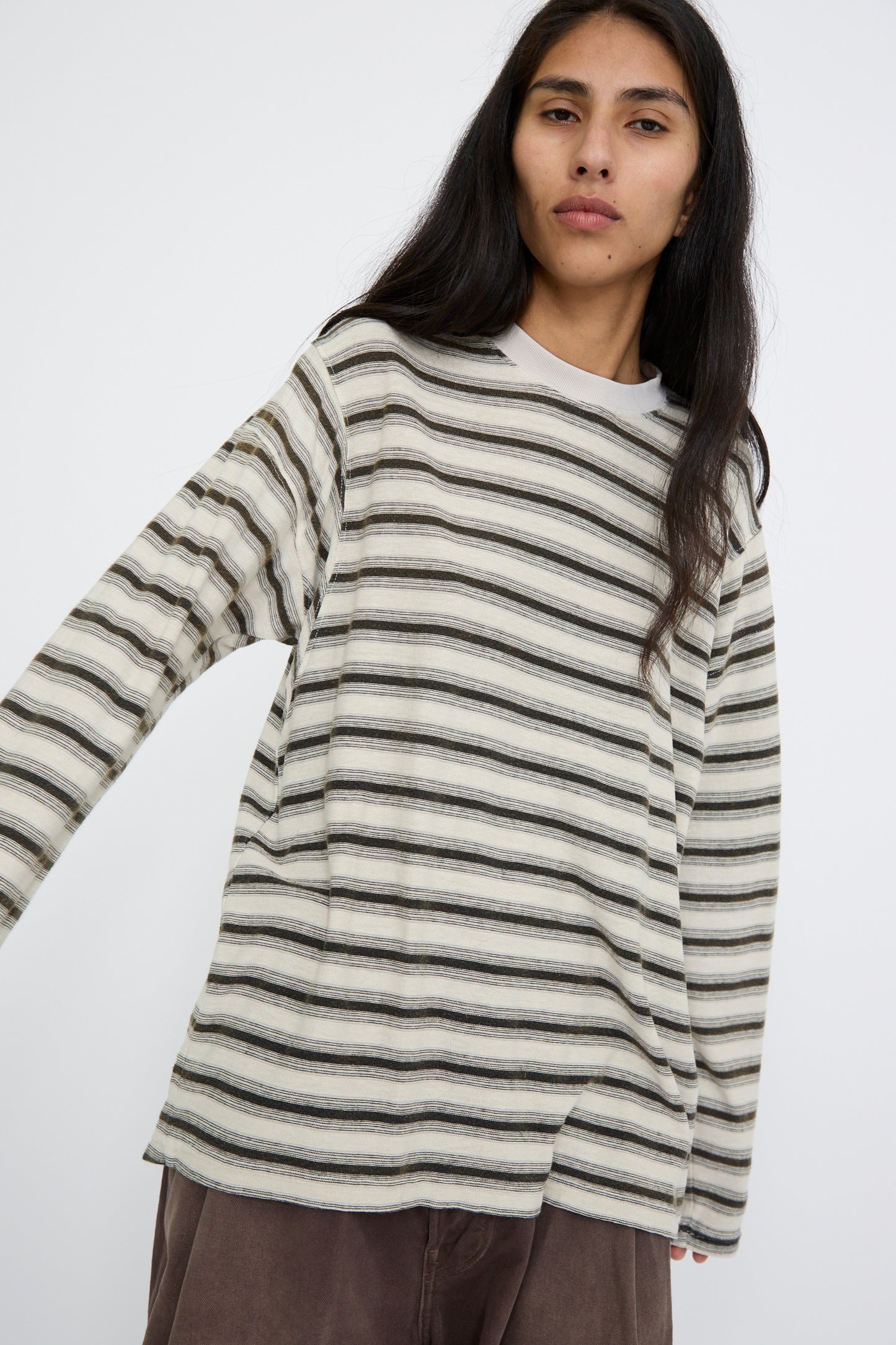 A person is standing against a plain background, wearing Dr. Collectors' relaxed-fit Hemp and Cotton Derrick Tee Stripes in Sunfaded Stone, paired with brown pants.