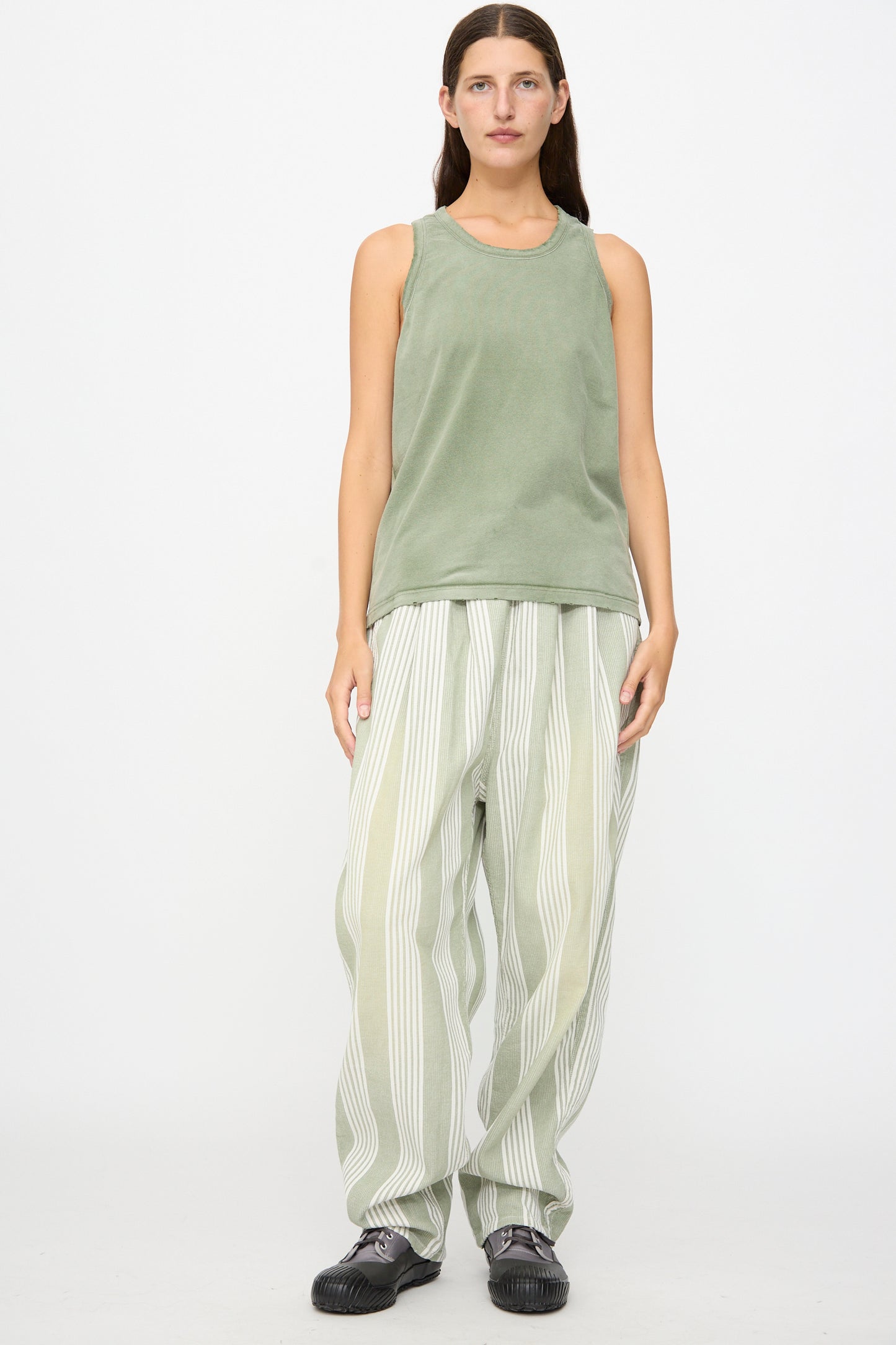 A person stands against a white background, wearing the Marcel Tank in Sunfaded Army Green by Dr. Collectors, paired with loose striped light green and white pants made with plant-based dyes, and dark shoes.
