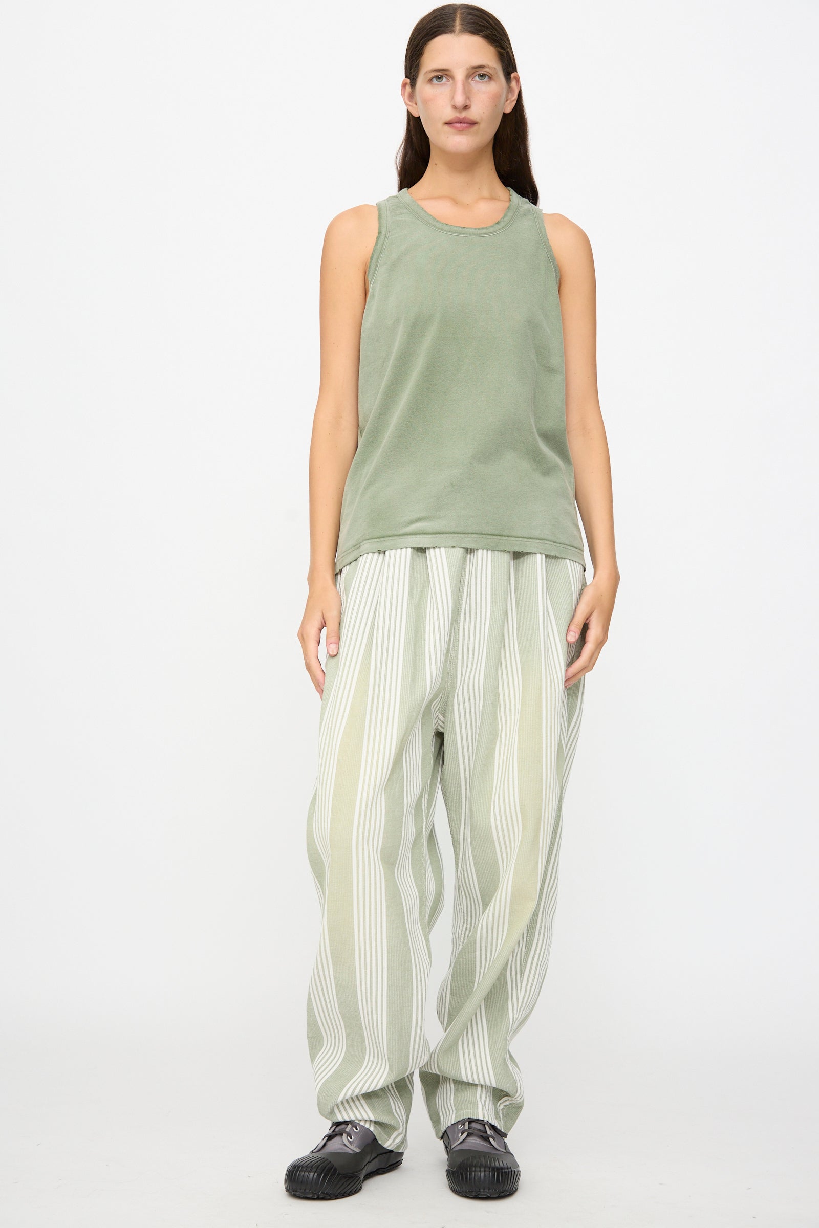 A person stands against a white background, wearing the Marcel Tank in Sunfaded Army Green by Dr. Collectors, paired with loose striped light green and white pants made with plant-based dyes, and dark shoes.