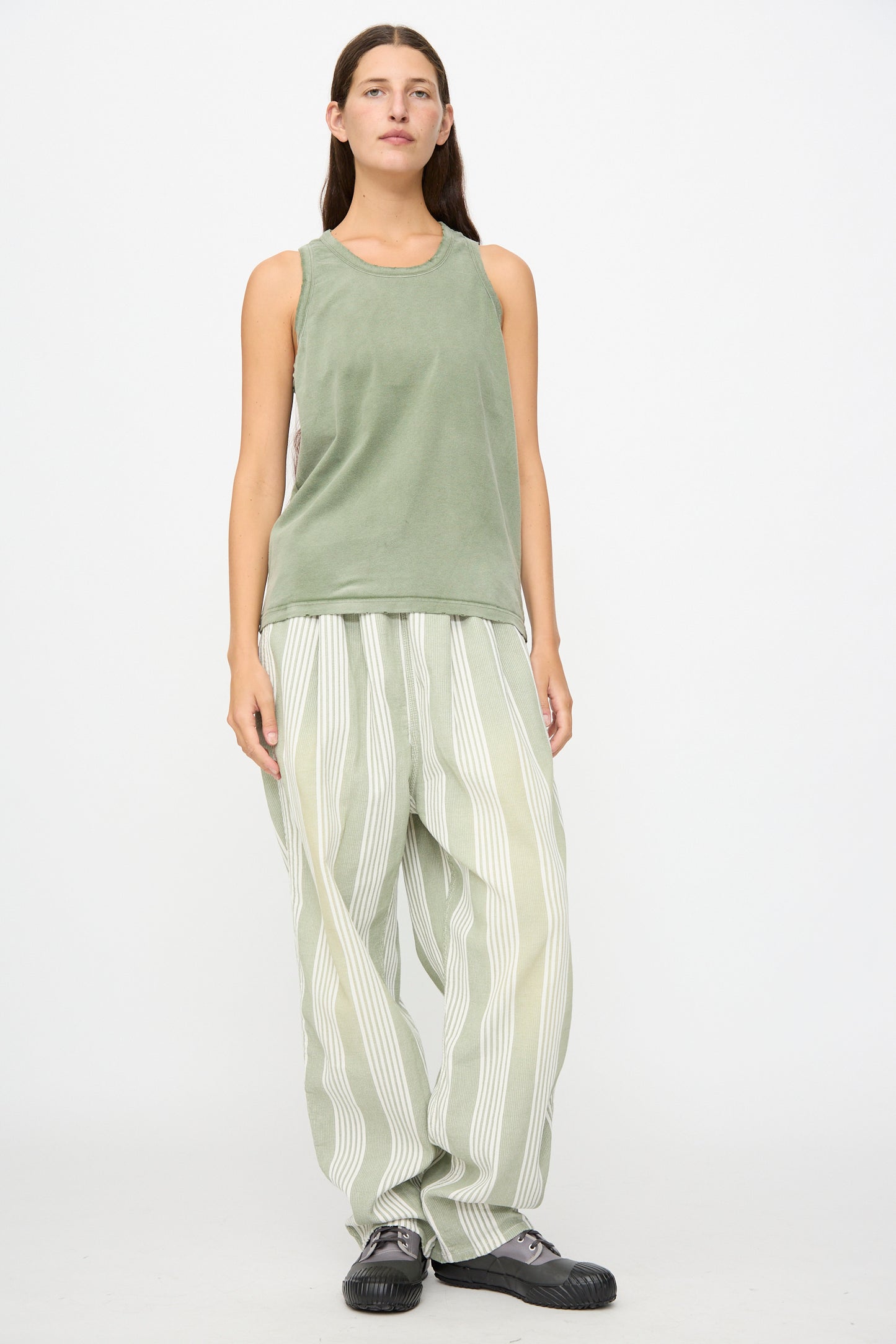 A person stands against a white background wearing the Marcel Tank in Sunfaded Army Green by Dr. Collectors, paired with light green and white striped loose pants dyed with plant-based dyes, and black shoes.