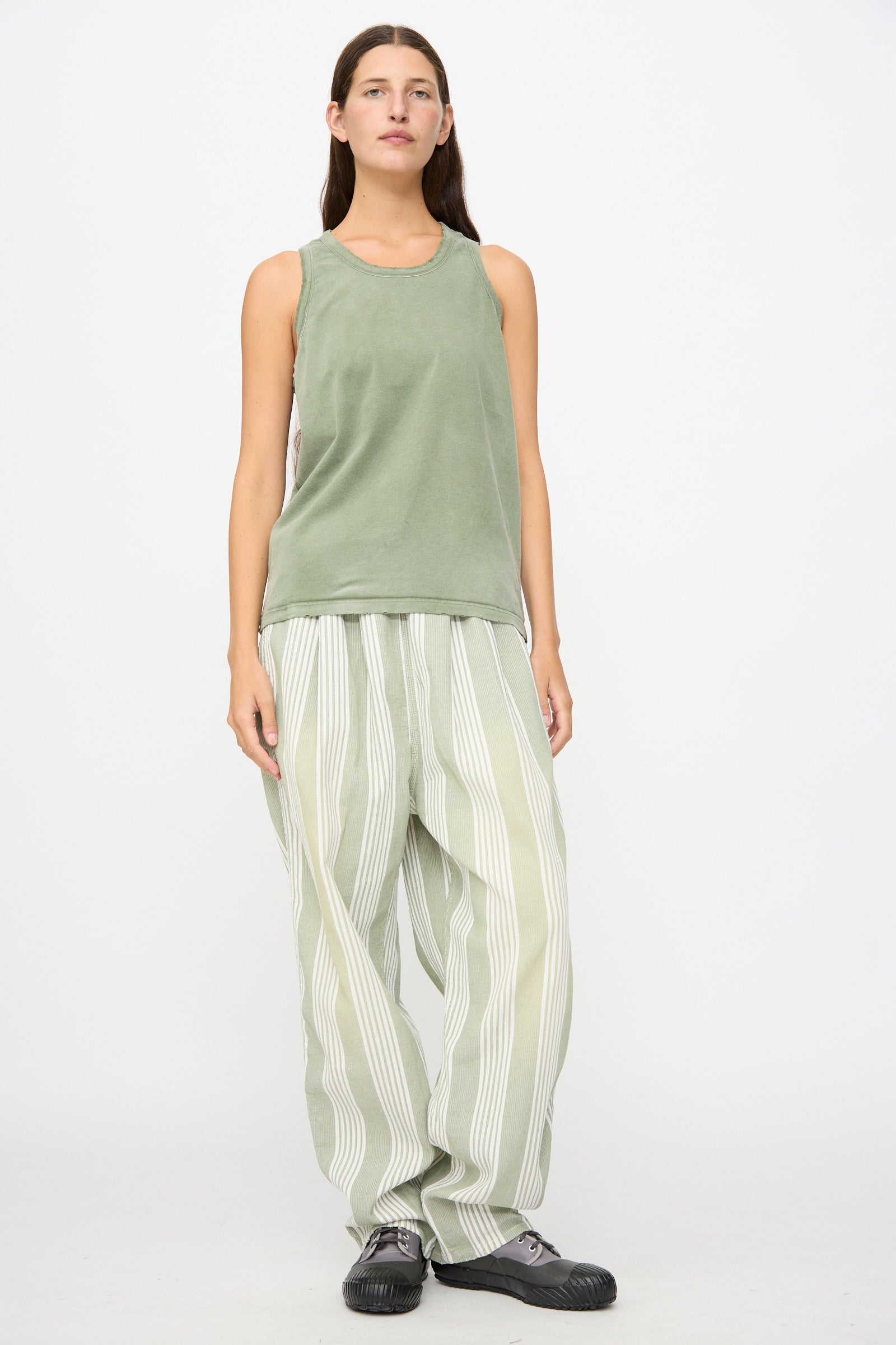 A person stands against a white background wearing the Marcel Tank in Sunfaded Army Green by Dr. Collectors, paired with light green and white striped loose pants dyed with plant-based dyes, and black shoes.