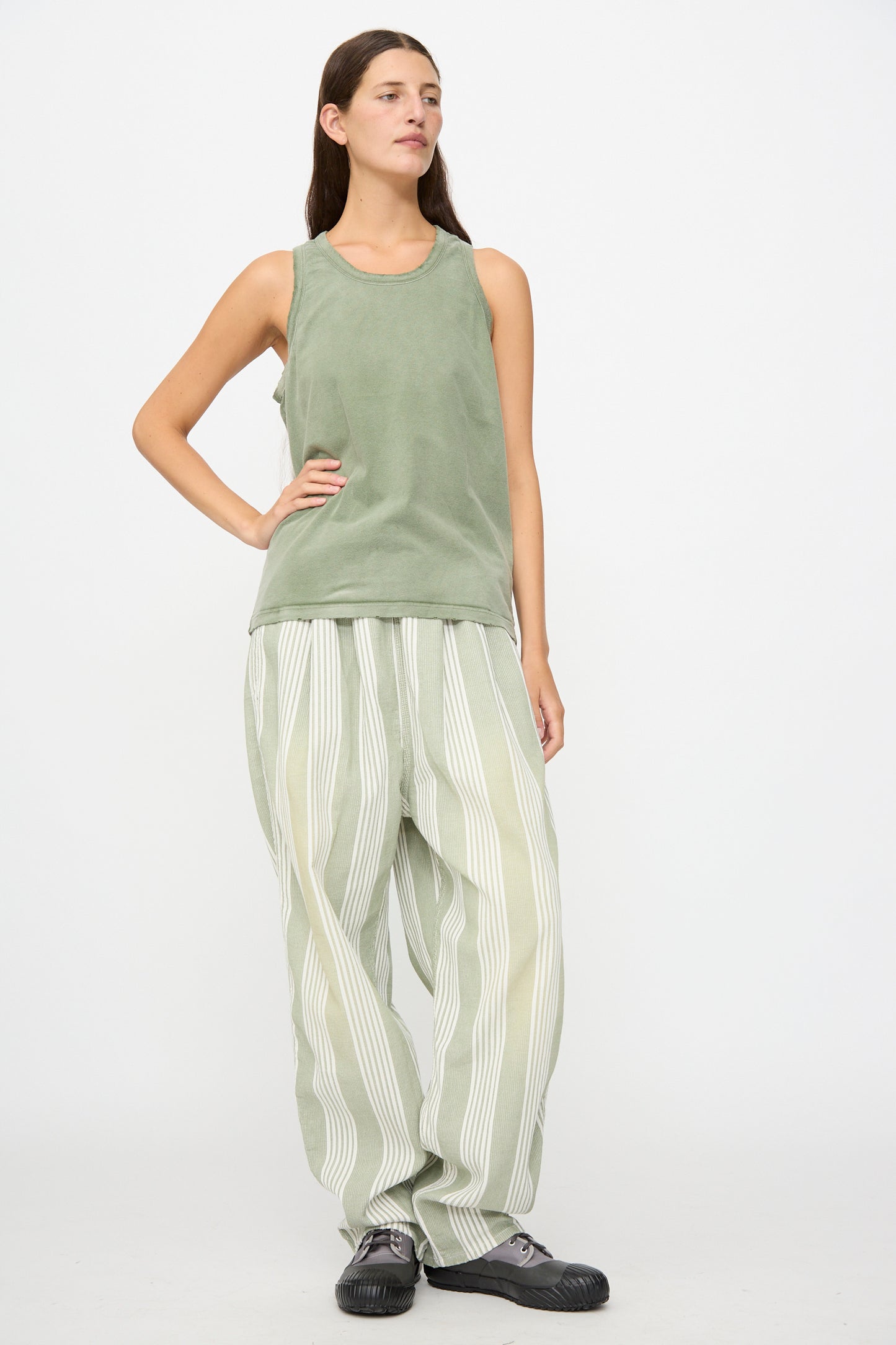 A person stands against a plain background, wearing the Marcel Tank in Sunfaded Army Green from Dr. Collectors, paired with loose striped pants dyed with plant-based dyes and black shoes; one hand is on their hip as they look slightly to the side.