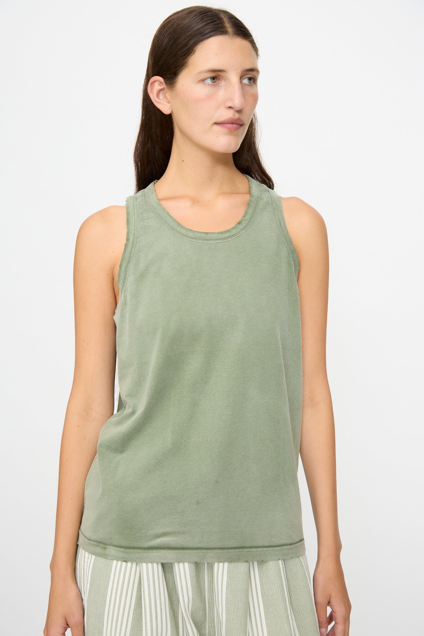 A woman with long brown hair wears the Marcel Tank in Sunfaded Army Green by Dr. Collectors, paired with striped pants, standing against a plain light background.