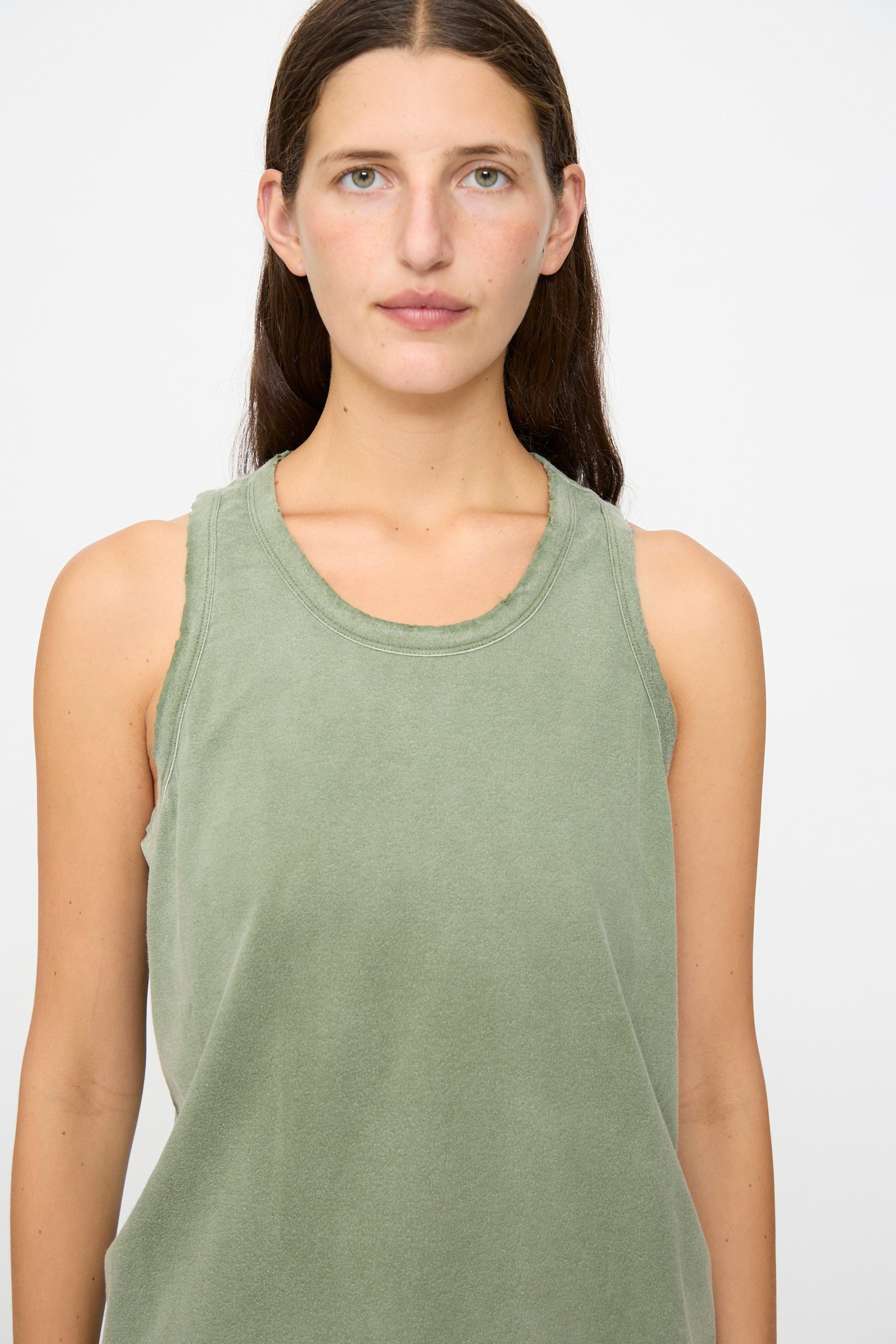 A person with long brown hair, wearing the Dr. Collectors Marcel Tank in Sunfaded Army Green—a sleeveless hemp top dyed with plant-based dyes—stands facing forward against a plain white background.
