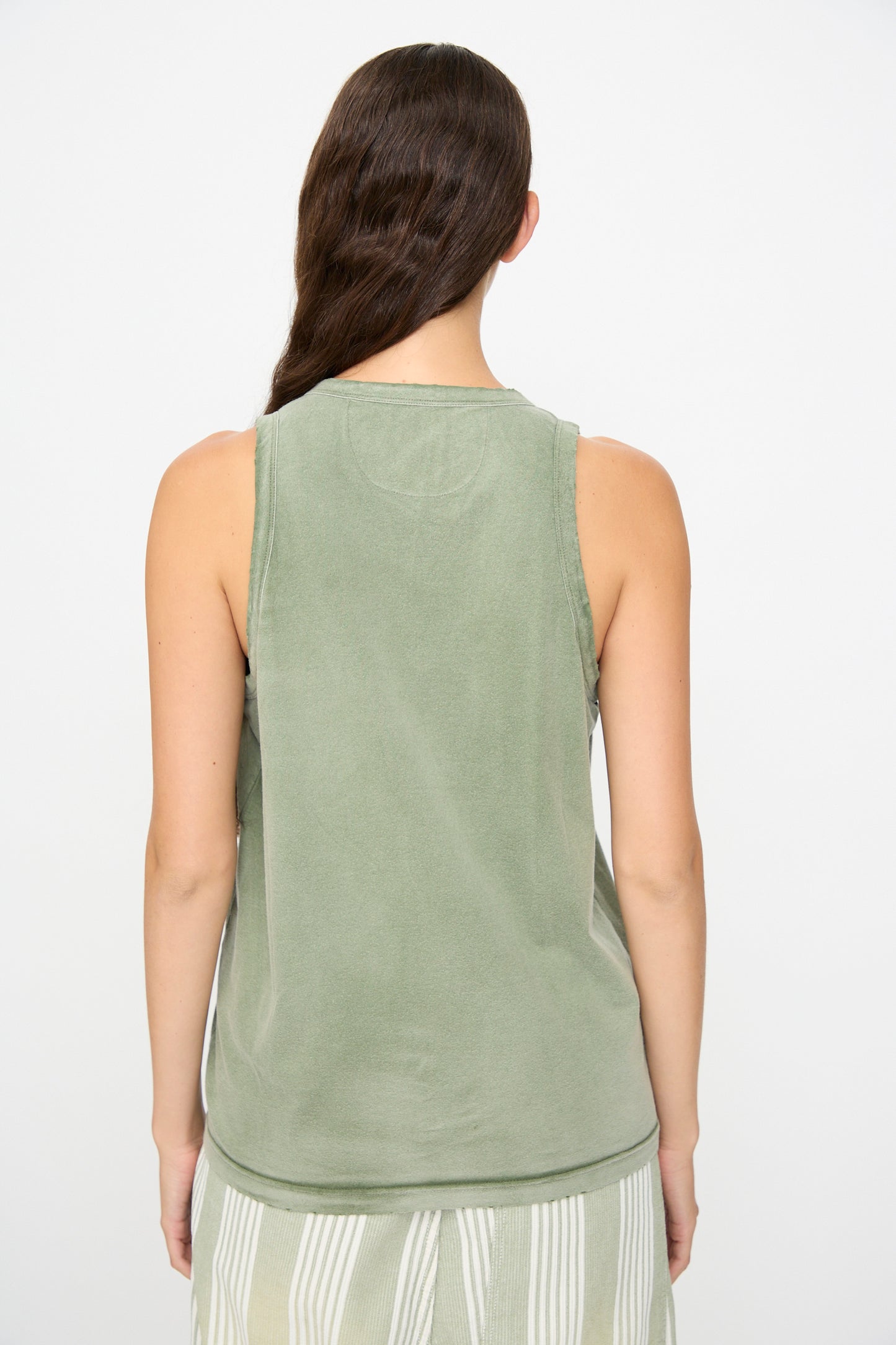 A person stands with their back to the camera, wearing the Marcel Tank in Sunfaded Army Green by Dr. Collectors—a sleeveless light green organic cotton top—paired with a skirt featuring vertical white and green stripes crafted from plant-based dyes.