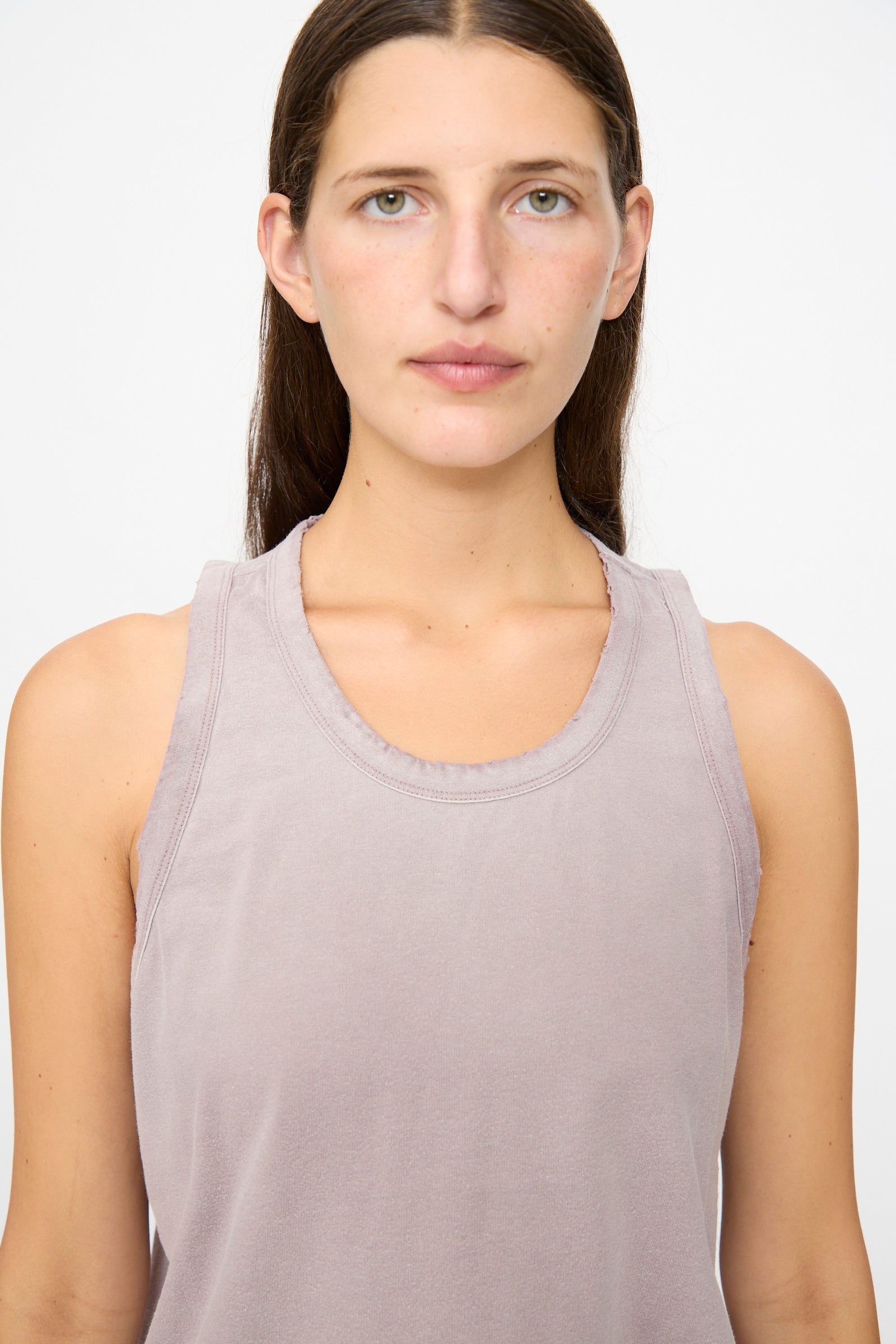 A person with long brown hair is standing facing forward against a plain white background, wearing the Dr. Collectors Marcel Tank in Sunfaded Dusty Orchid—a sleeveless, light purple organic cotton top colored with plant-based dyes.