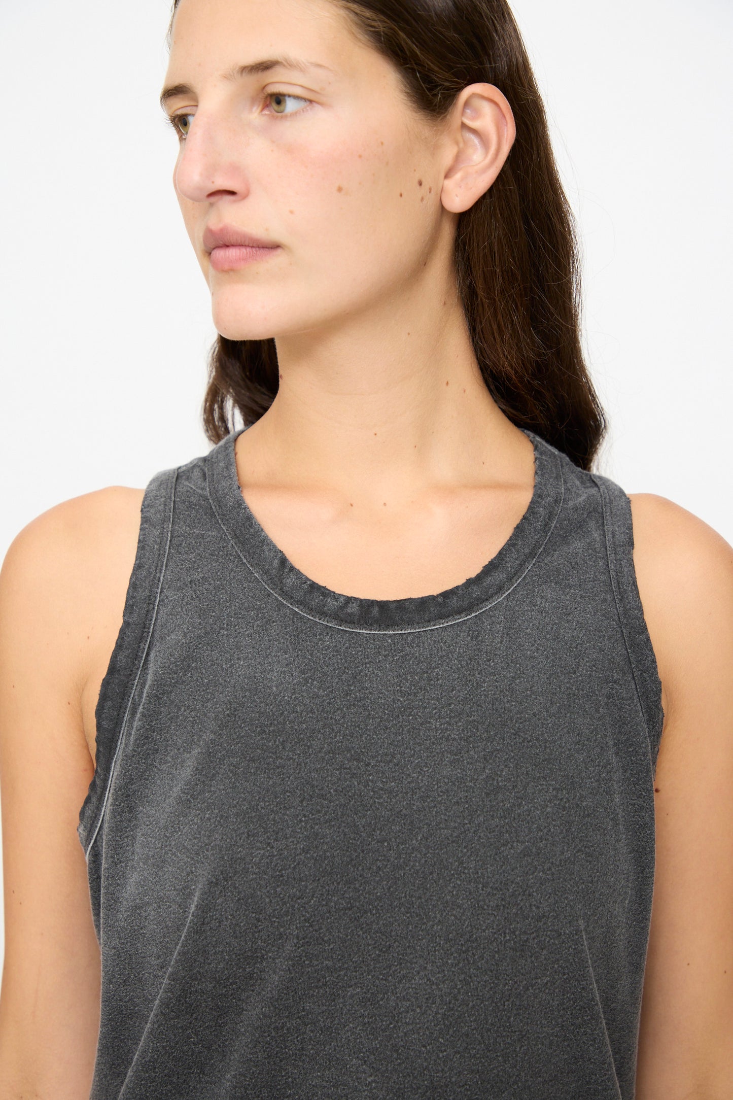 A person with long dark hair, wearing the Marcel Tank in Sunfaded Smoke Black by Dr. Collectors—a sleeveless hemp top dyed with natural plant-based dyes—looks to the left against a plain light background.