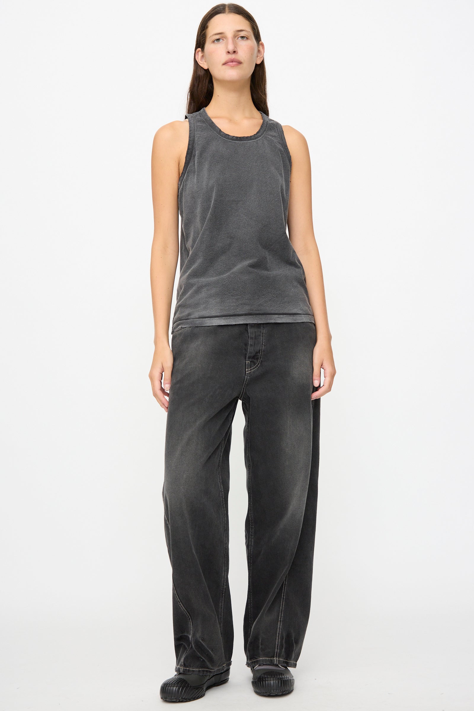 A person stands against a white background wearing a sleeveless gray top, loose-fitting dark jeans, and black shoes.