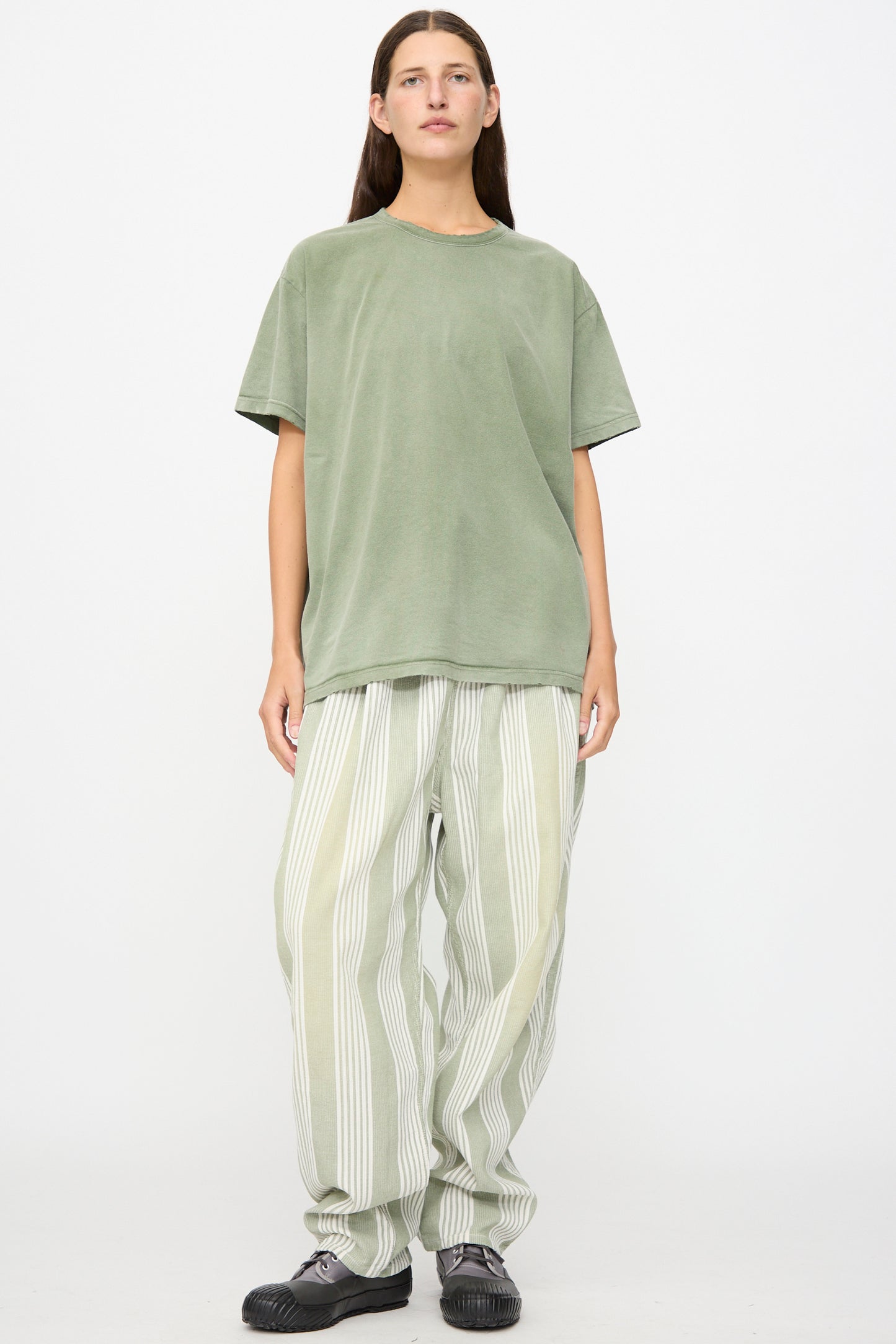 A person stands against a white background wearing the Dr. Collectors Model T in Sunfaded Army Green—a loose-fit tee crafted from recycled cotton—paired with wide striped light-colored pants and black shoes.