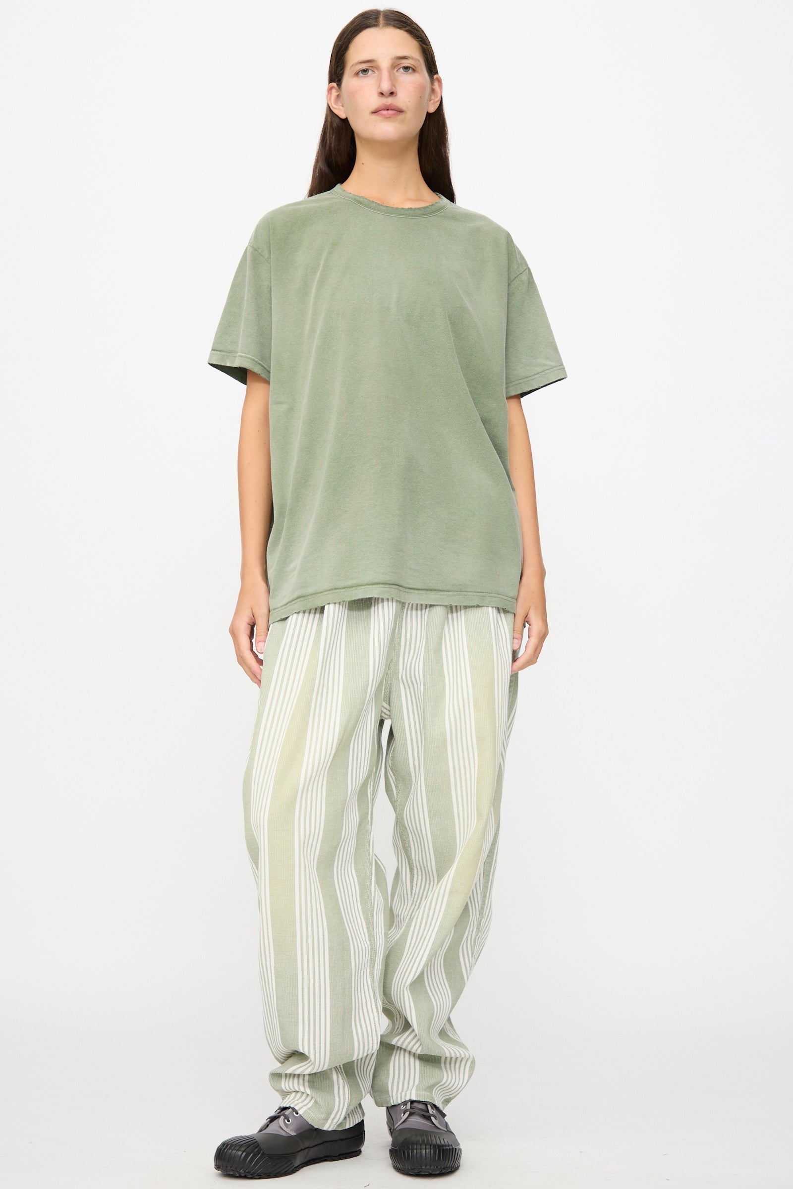 A person stands against a white background wearing the Dr. Collectors Model T in Sunfaded Army Green—a loose-fit tee crafted from recycled cotton—paired with wide striped light-colored pants and black shoes.