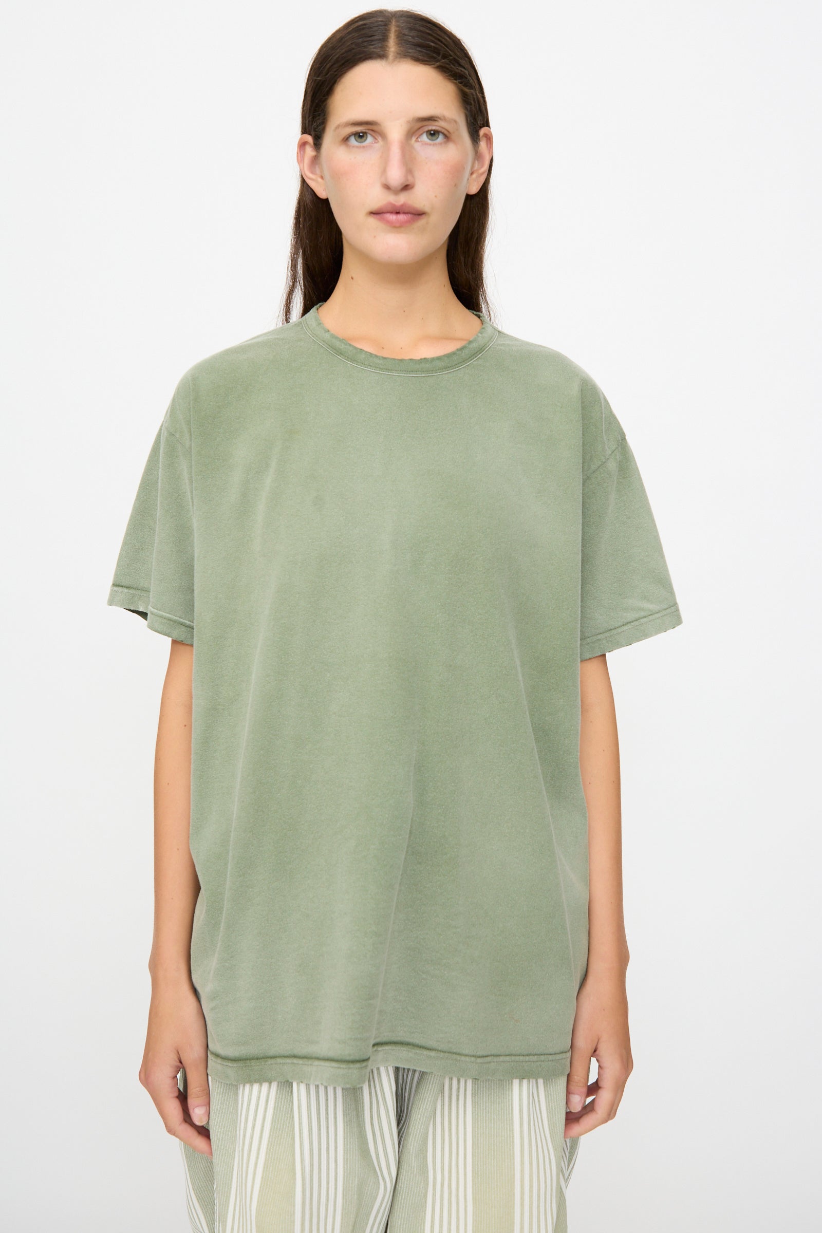 A person with a neutral expression and long brown hair stands facing forward against a plain white background, wearing the Dr. Collectors Model T in Sunfaded Army Green—an oversized, loose fit tee crafted from recycled cotton—paired with striped pants.