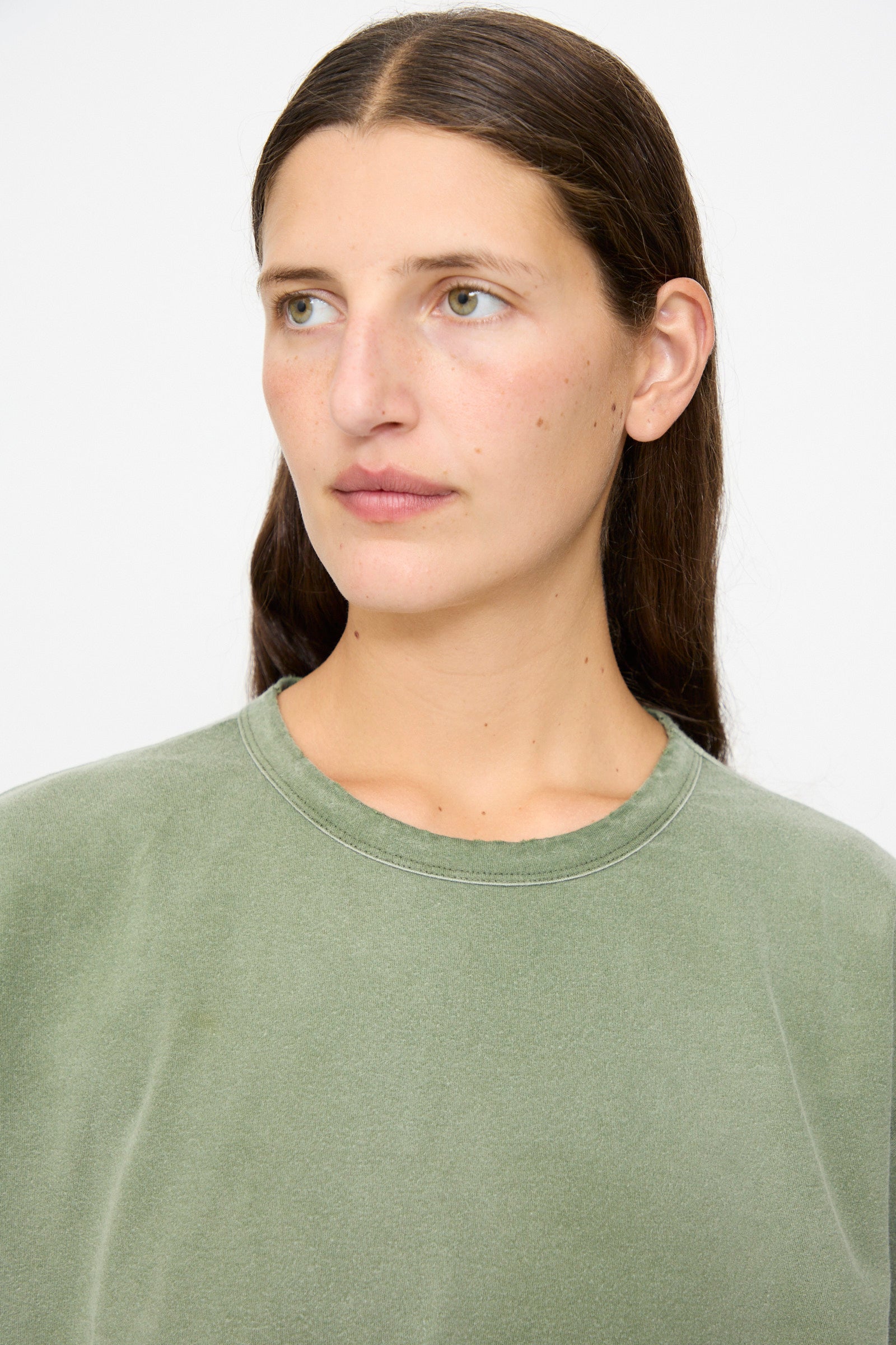 A person with long brown hair, wearing the Dr. Collectors Model T in Sunfaded Army Green crew neck shirt made from recycled cotton, looks slightly to the side against a plain light background.