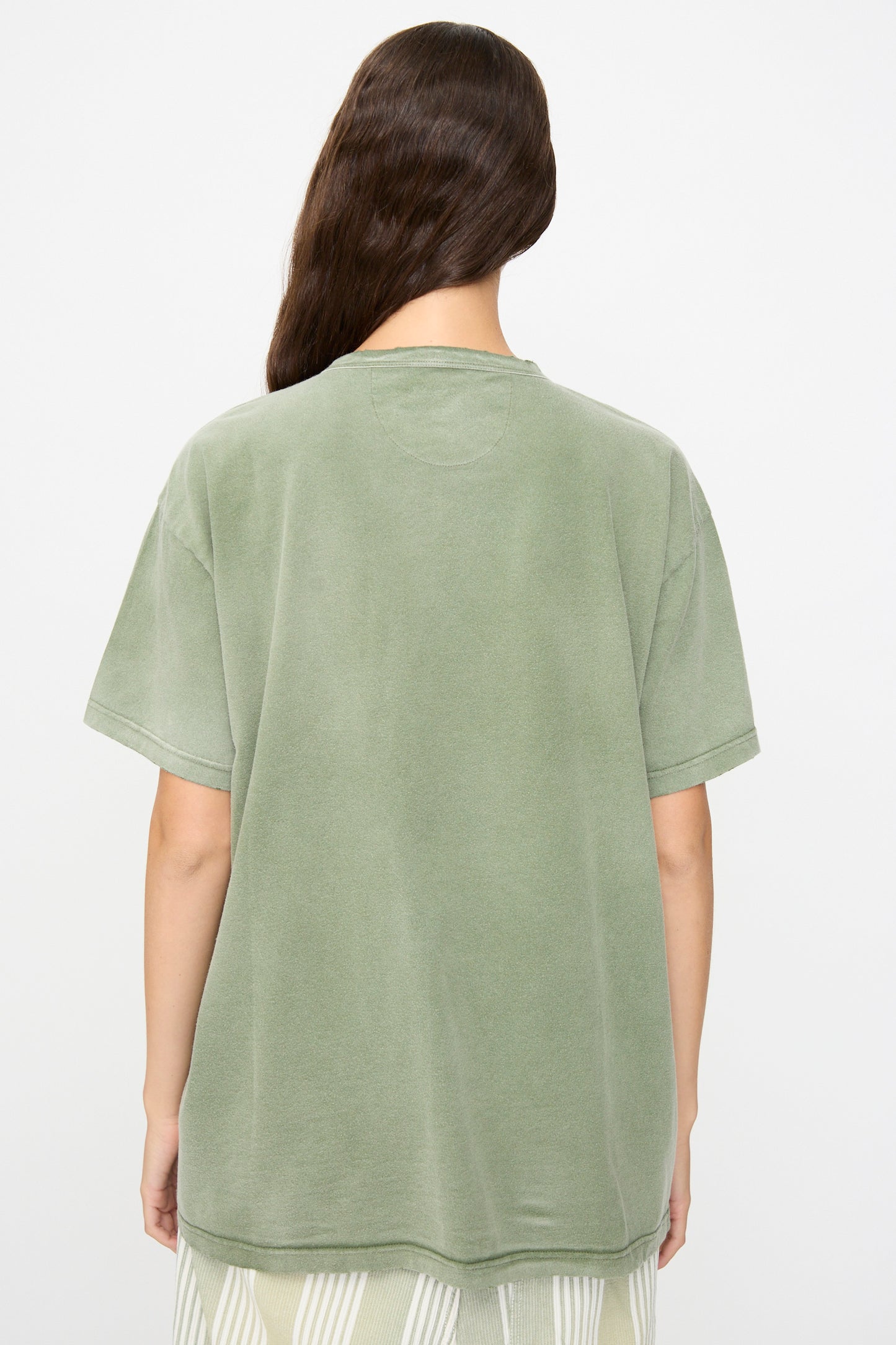 A person stands with their back to the camera, wearing Dr. Collectors’ Model T in Sunfaded Army Green—a loose-fitting, short-sleeved tee crafted from recycled cotton—paired with a light-colored striped skirt for a relaxed look with subtle vintage appeal.