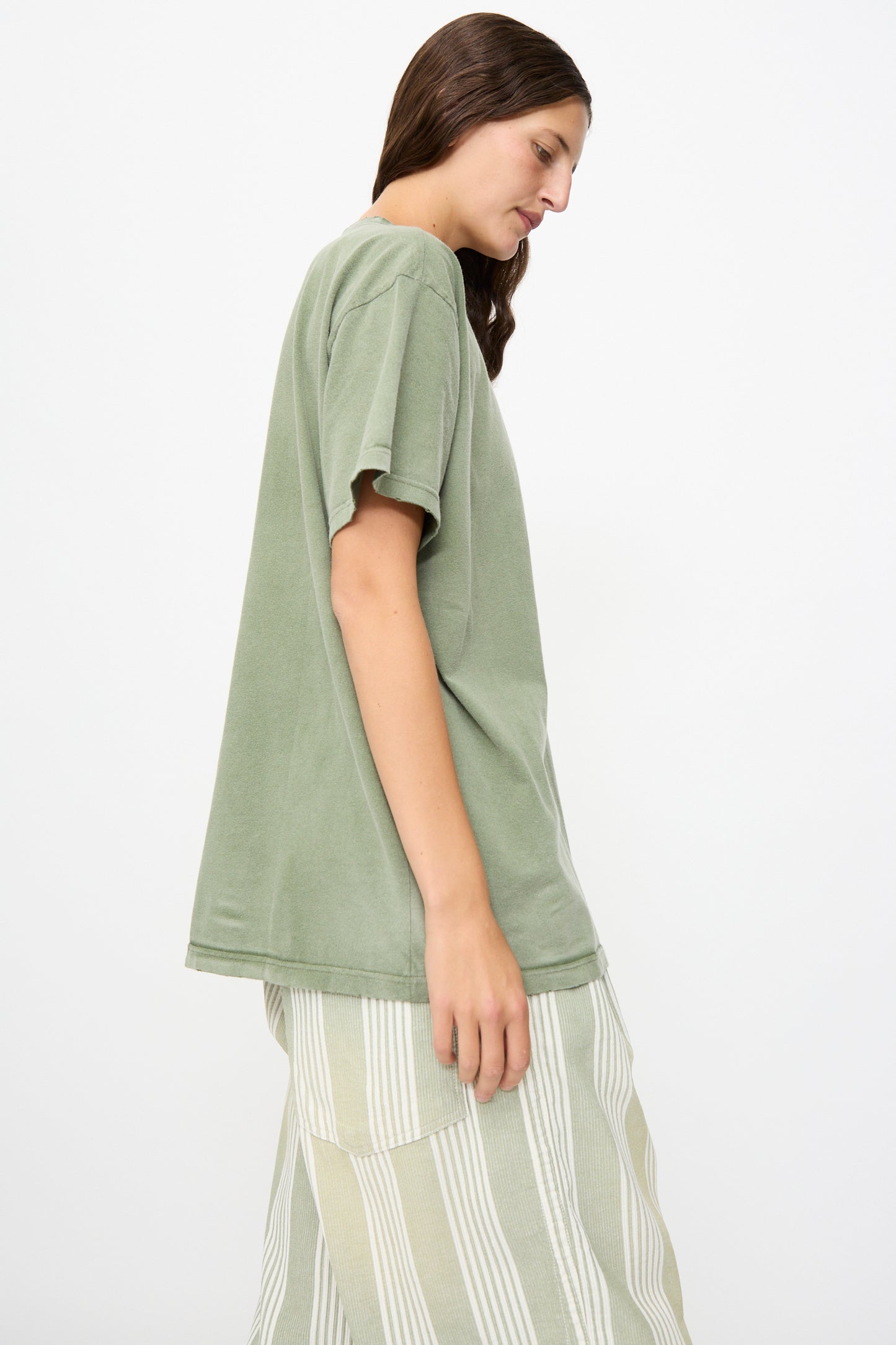 A person stands sideways against a plain white background, showcasing a relaxed, vintage appearance in Dr. Collectors’ Model T tee in Sunfaded Army Green—crafted from recycled cotton and styled with light-colored, striped pants.