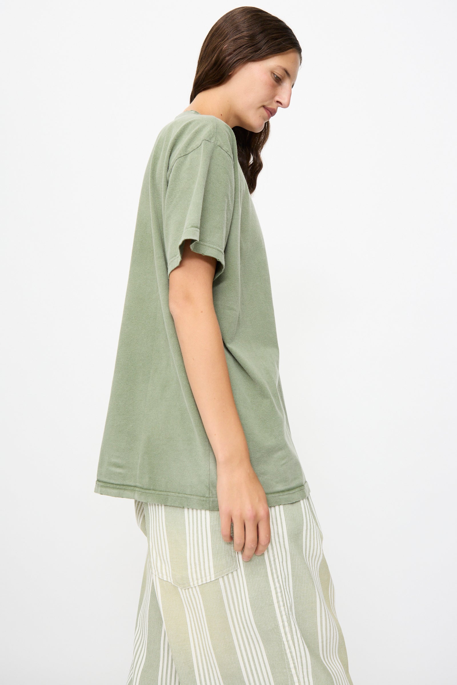 A person stands sideways against a plain white background, showcasing a relaxed, vintage appearance in Dr. Collectors’ Model T tee in Sunfaded Army Green—crafted from recycled cotton and styled with light-colored, striped pants.