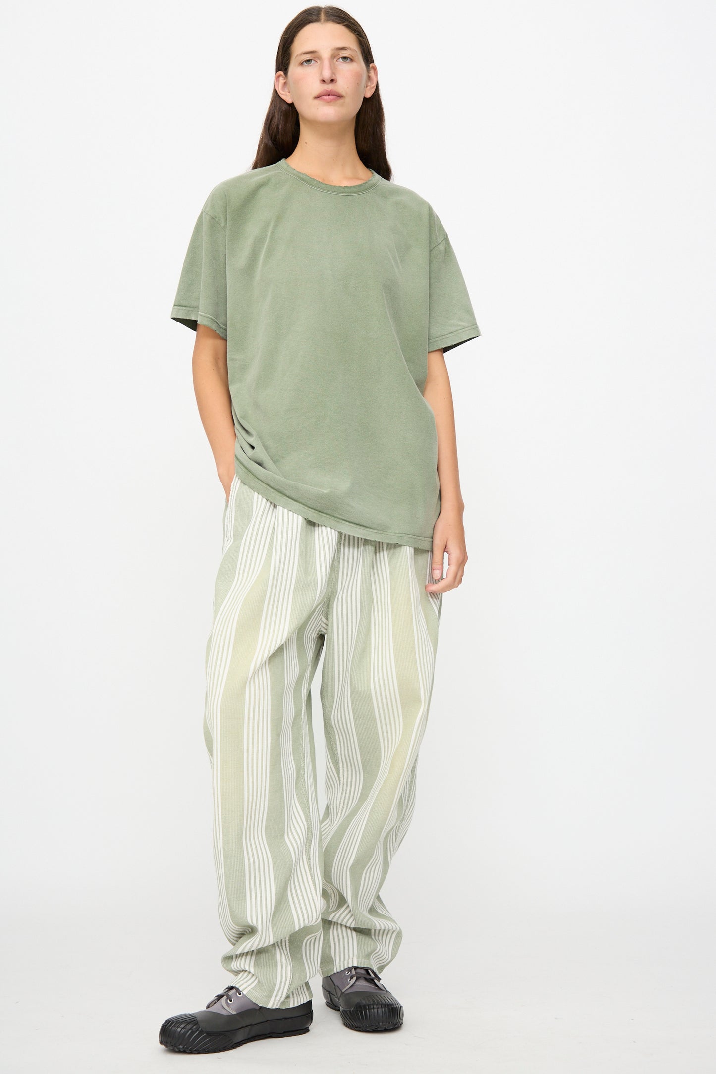A person stands against a plain background, wearing the Dr. Collectors Model T in Sunfaded Army Green—a loose fit tee—paired with striped light green and white pants made from recycled cotton and black shoes.