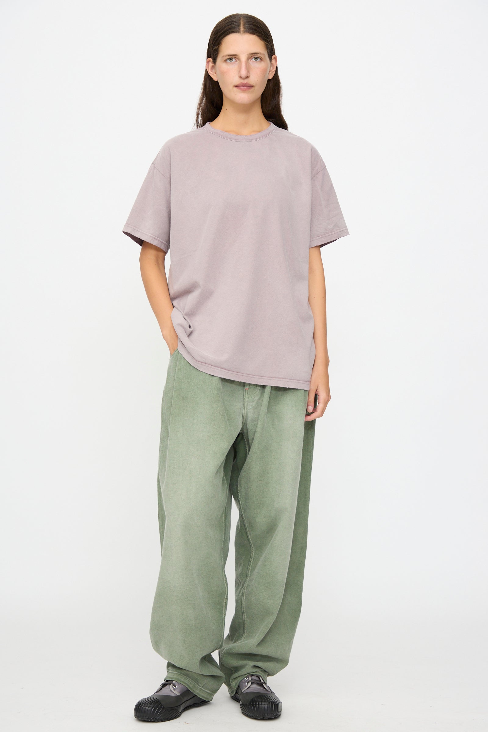A person stands against a plain background, wearing the Dr. Collectors Model T in Sunfaded Dusty Orchid—a loose fit tee crafted from recycled cotton—paired with baggy sage green pants and black sneakers, one hand casually in their pocket. 