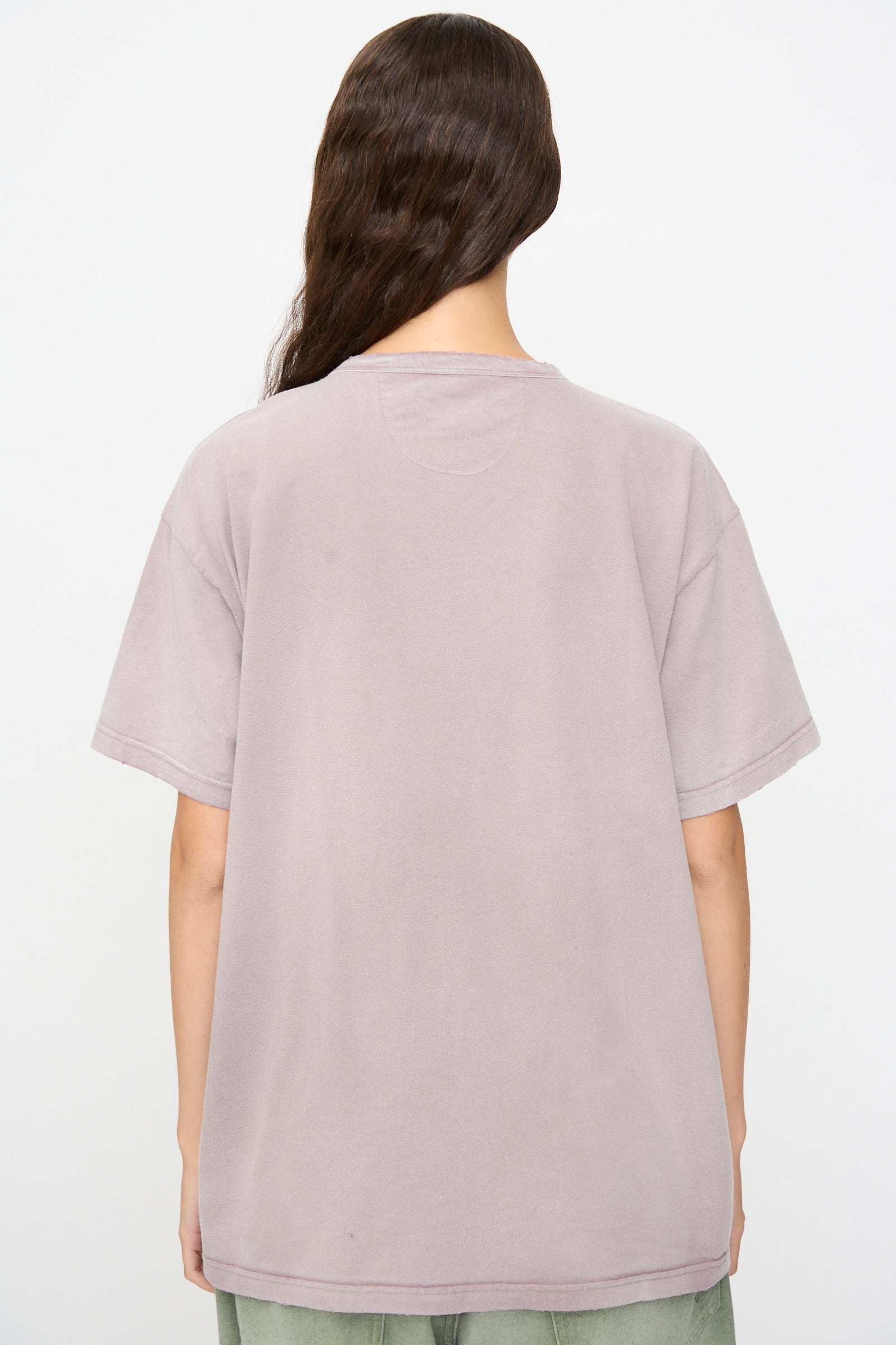 Person standing with their back to the camera, wearing the Dr. Collectors Model T in Sunfaded Dusty Orchid—a loose fit tee crafted from recycled cotton in light mauve—paired with light green pants, lending a subtle vintage appearance against a plain white background.