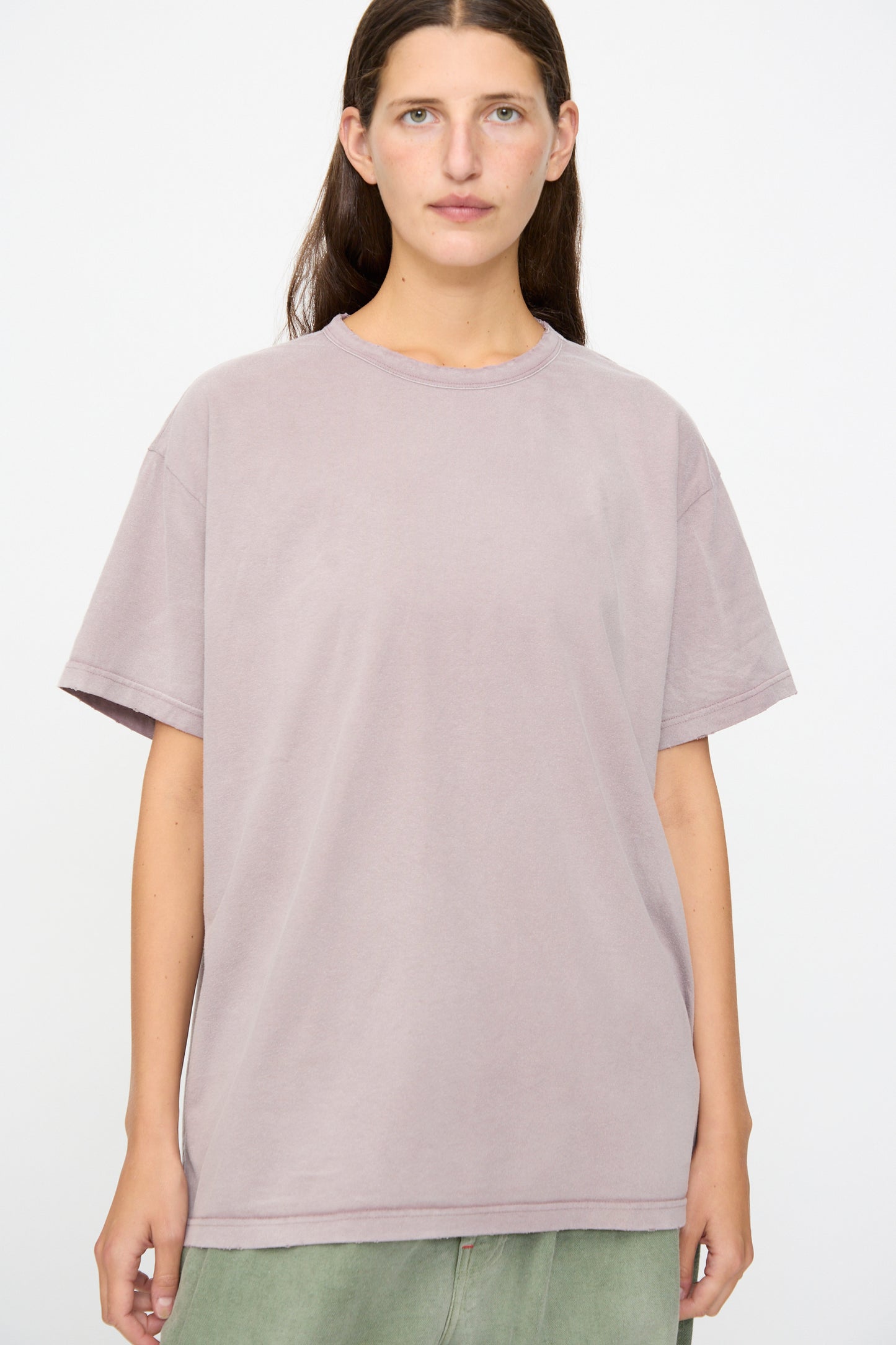 A person stands against a plain background, wearing Dr. Collectors’ Model T in Sunfaded Dusty Orchid—a loose-fitting tee crafted from recycled cotton —paired with green pants and facing forward with a neutral expression.