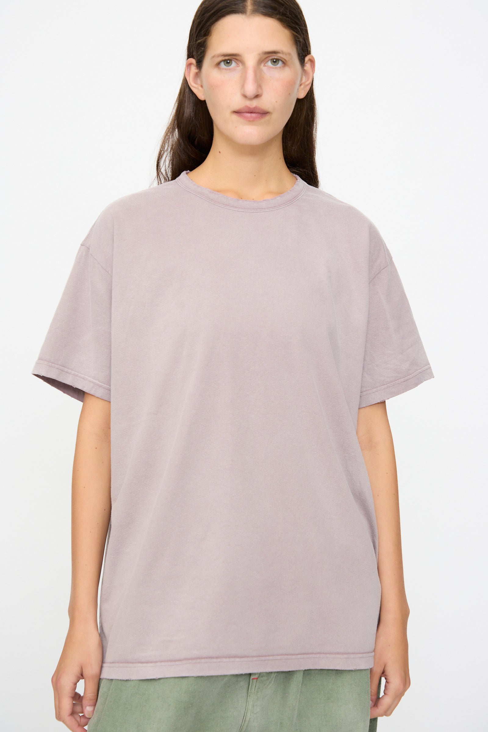 A person stands against a plain background, wearing Dr. Collectors’ Model T in Sunfaded Dusty Orchid—a loose-fitting tee crafted from recycled cotton —paired with green pants and facing forward with a neutral expression.