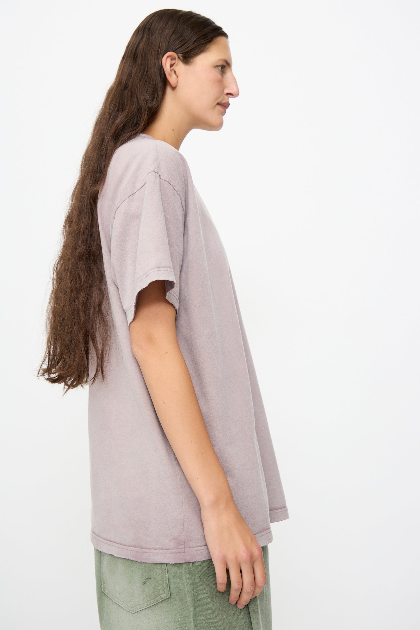 A person with long brown hair is shown in profile against a plain white background, wearing the Dr. Collectors Model T in Sunfaded Dusty Orchid—a loose fit, light purple t-shirt made from recycled cotton—and green pants.