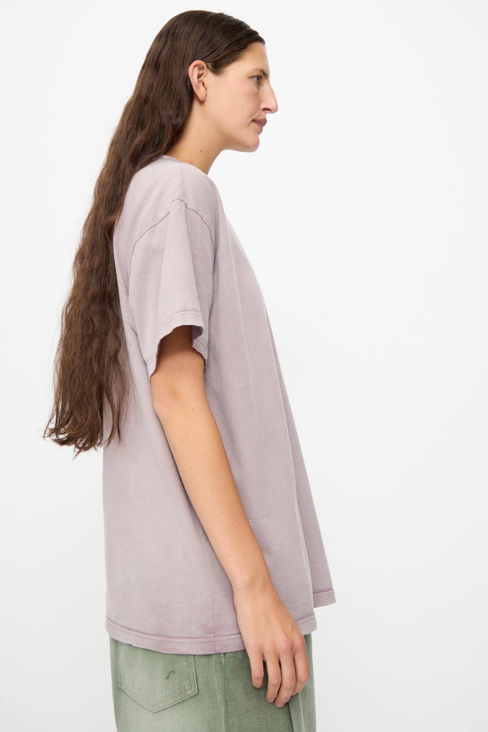 A person with long brown hair is shown in profile against a plain white background, wearing the Dr. Collectors Model T in Sunfaded Dusty Orchid—a loose fit, light purple t-shirt made from recycled cotton—and green pants.