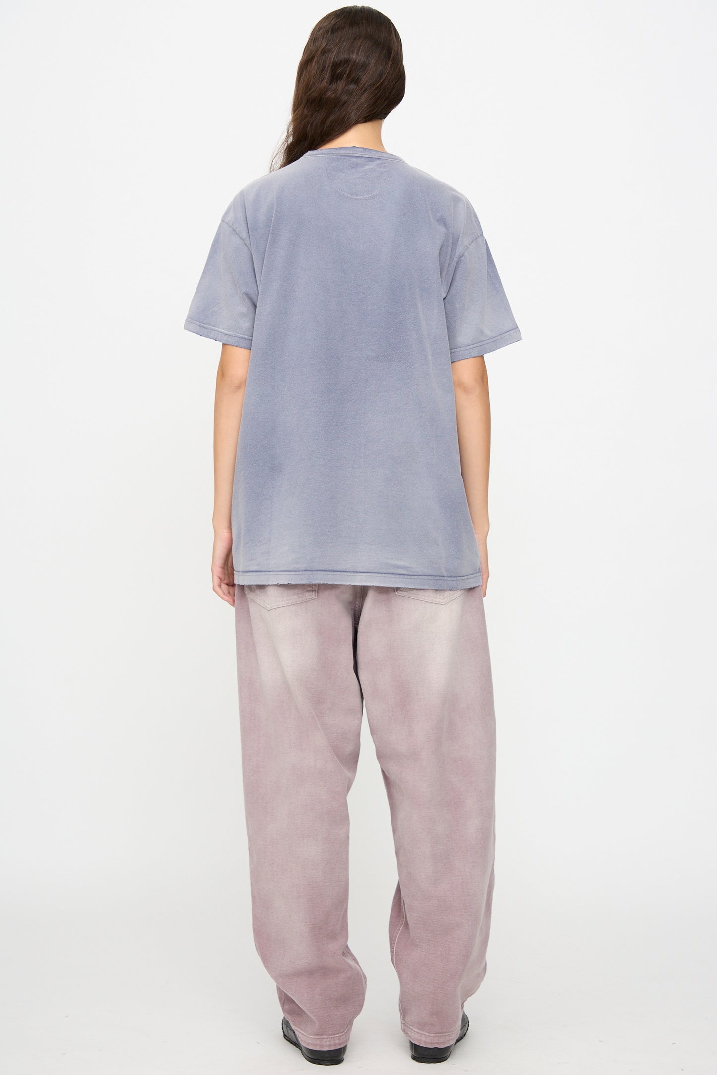 A person stands with their back to the camera, wearing Dr. Collectors’ Model T in Sunfaded Navy—a loose fit tee—paired with baggy light purple jeans, set against a plain white background.