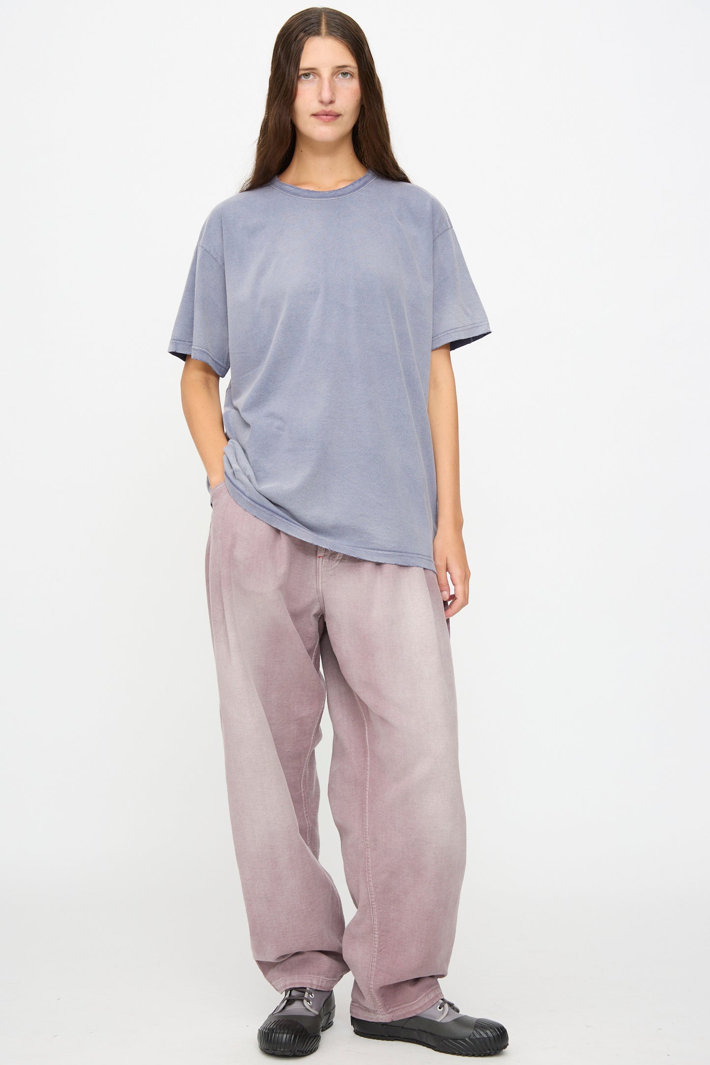 A person stands against a white background wearing the Dr. Collectors Model T in Sunfaded Navy, paired with baggy faded pink pants and black shoes, with one hand in their pocket.