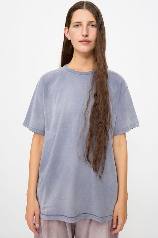 A person with long brown hair wearing Dr. Collectors’ Model T in Sunfaded Navy—a loose fit tee made from vintage cotton t-shirt fabric—and light pants stands against a plain white background.