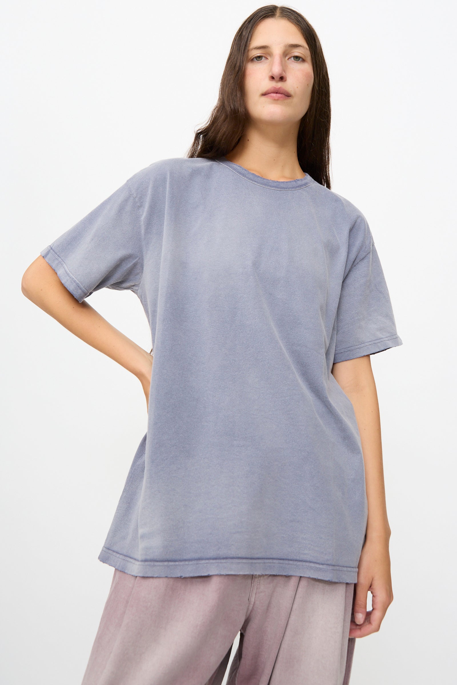 A woman with long brown hair wears Dr. Collectors’ Model T in Sunfaded Navy—a loose fit vintage cotton t-shirt—paired with light pink pants, standing against a plain white background.