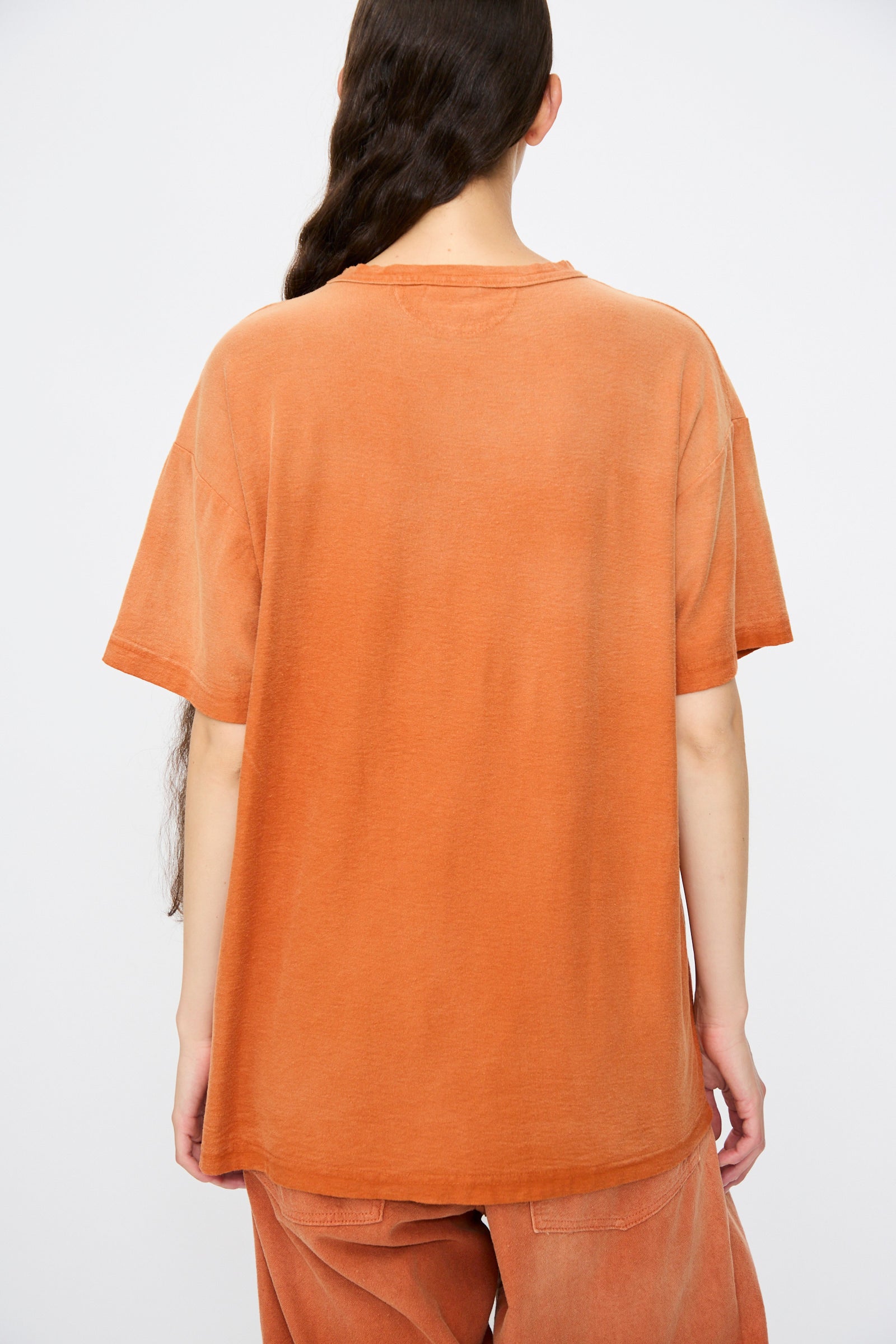 A person stands with their back to the camera, wearing Dr. Collectors' Model T. Sunfaded in Rust—a loose fit tee and matching orange pants made from recycled cotton for a cozy, vintage look.